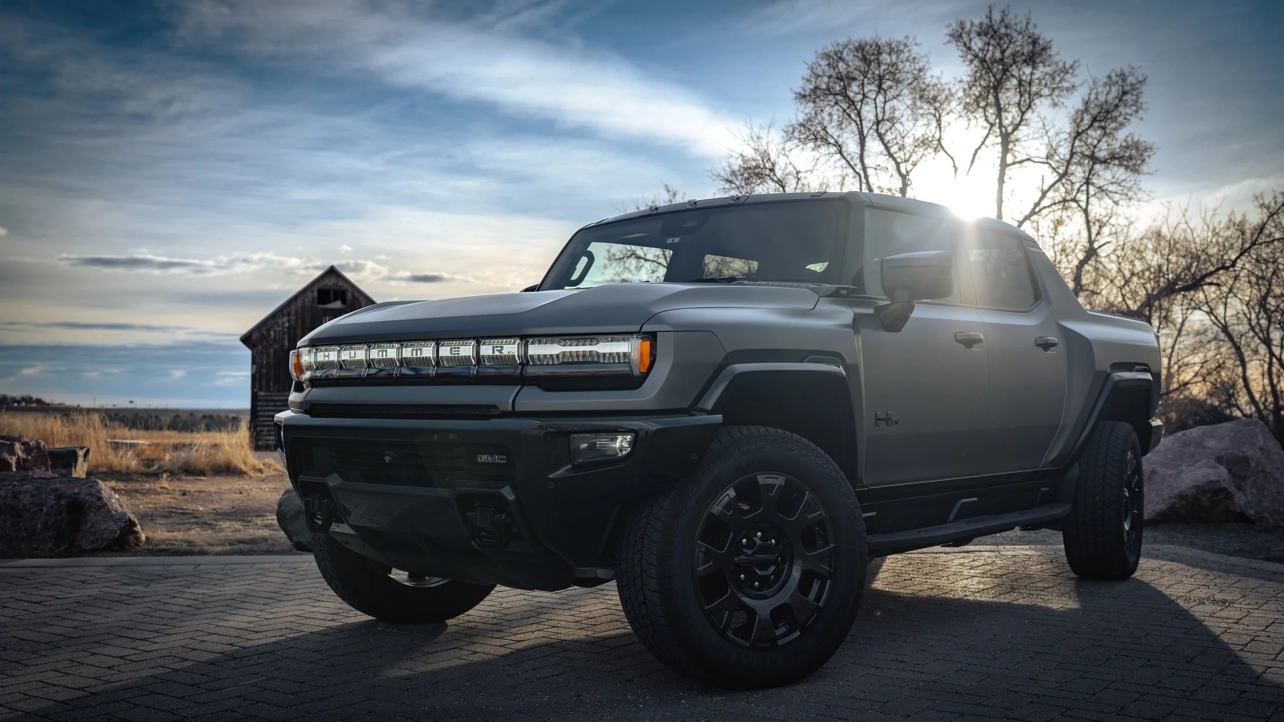 A black GMC Hummer EV truck parked outdoors on a stone pavement with dry grass, rocks, leafless trees, and a barn under a partly cloudy sky with the sun setting or rising in the background.