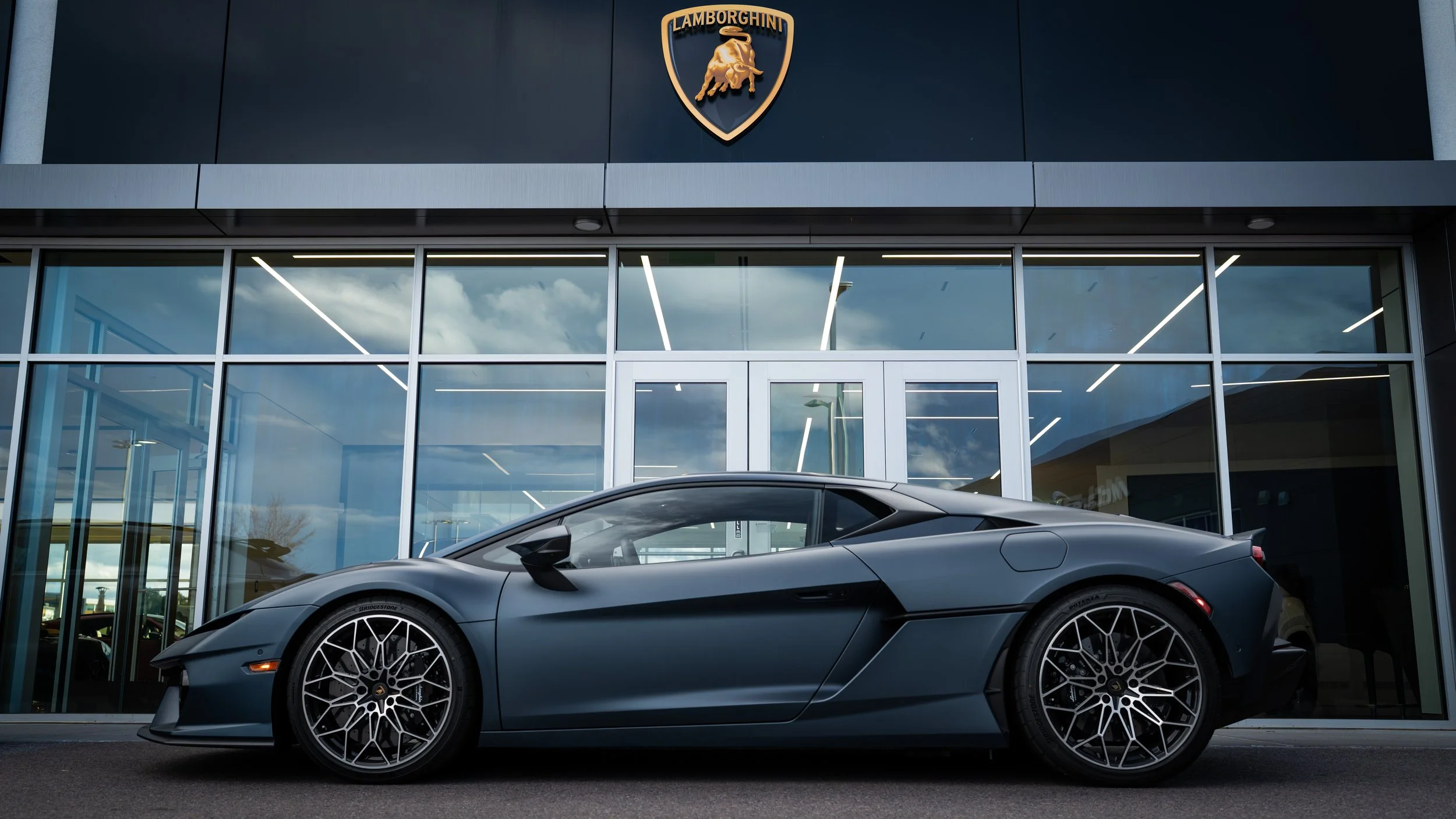 A sleek gray Lamborghini sports car parked in front of a modern car dealership with large glass windows and a Lamborghini logo on the building. The sky is partly cloudy and the reflection of the sky can be seen in the windows.