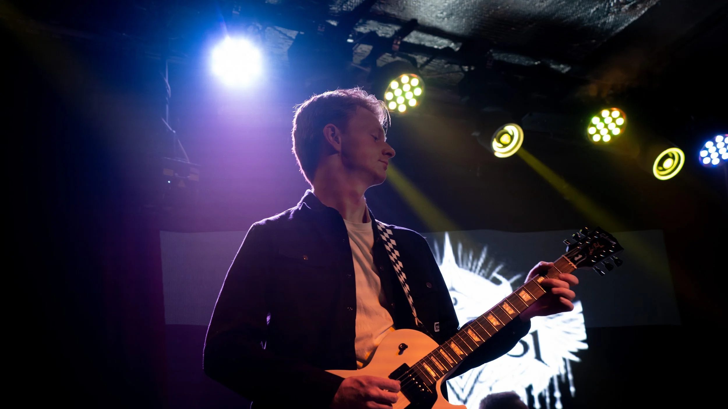 A young man playing an electric guitar on stage under colorful stage lights.