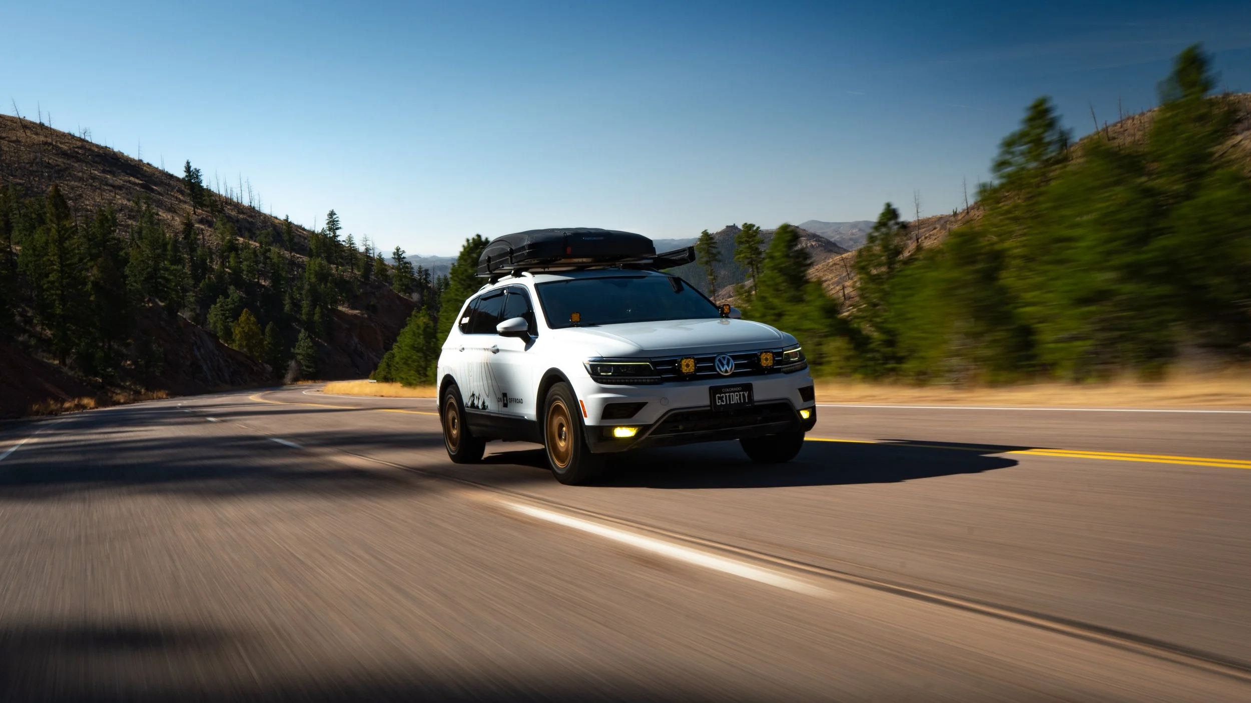 A white Volkswagen SUV driving on a winding mountain road with a cargo box on top, surrounded by pine trees and hills under a clear blue sky.