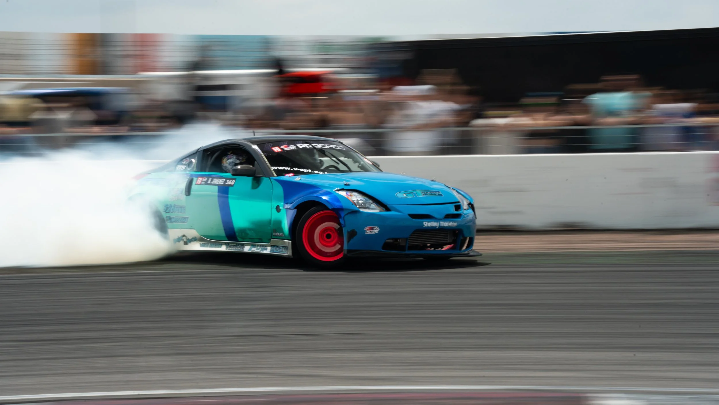 A blue and teal race car drifting on a track, producing tire smoke, with a blurred background of spectators and structures.