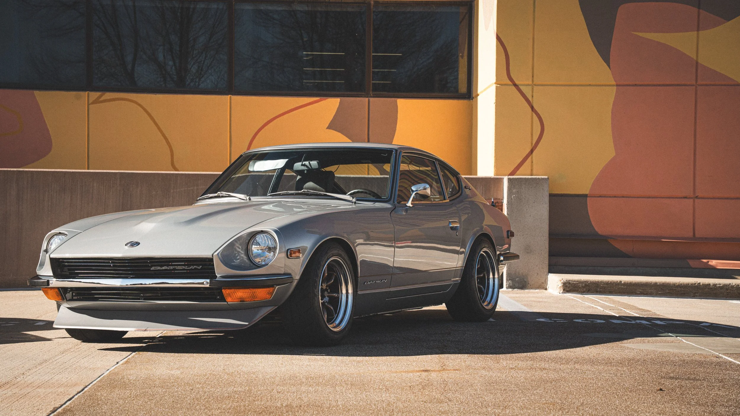 A classic silver Datsun 240Z parked in a lot in front of a modern building with a colorful abstract mural, with leafless trees reflected in the upper window.