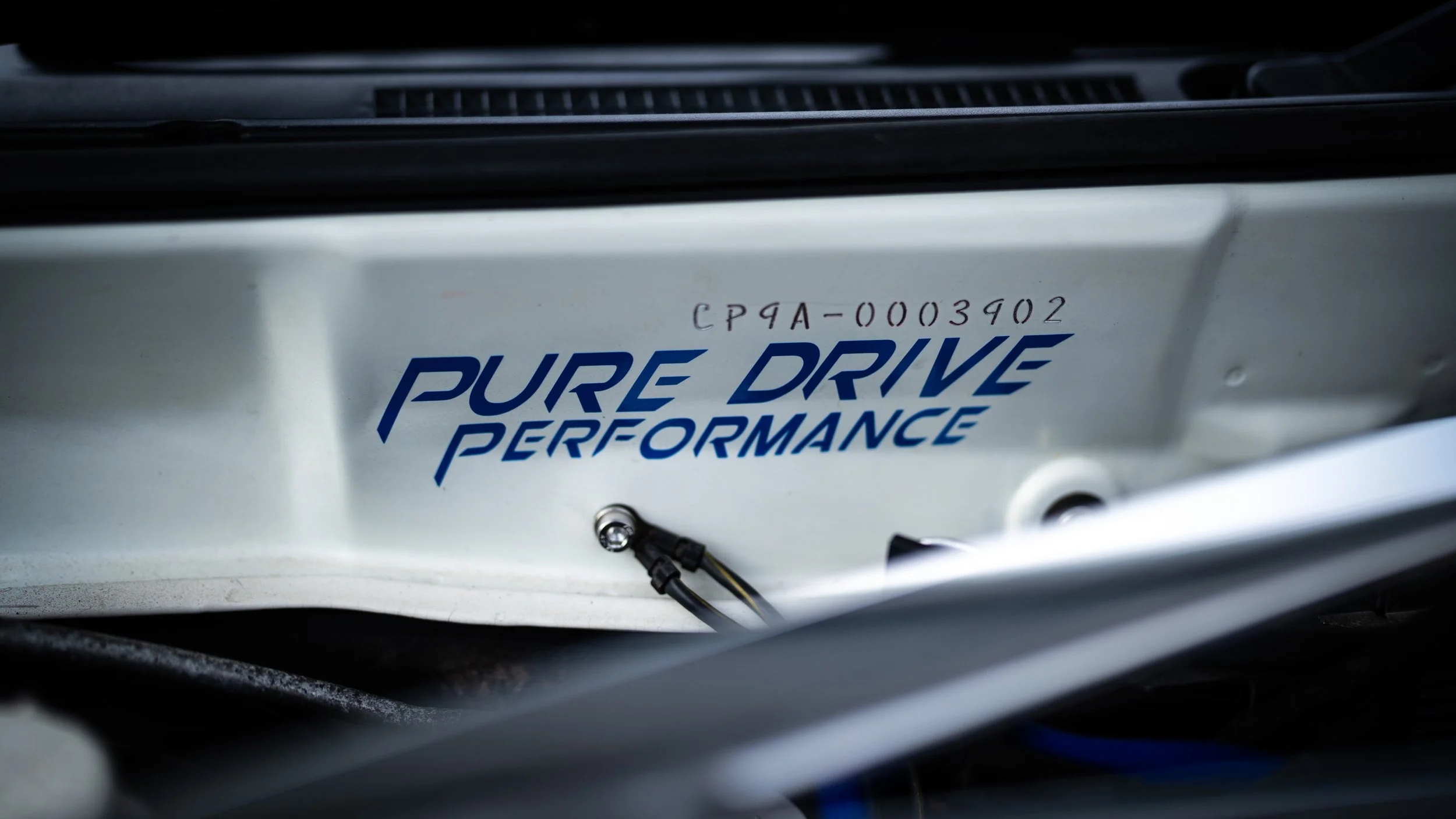 Close-up of a car component with a white surface, blue text reading 'Pure Drive Performance,' and a handwritten code 'CP9A-0003902' above it.