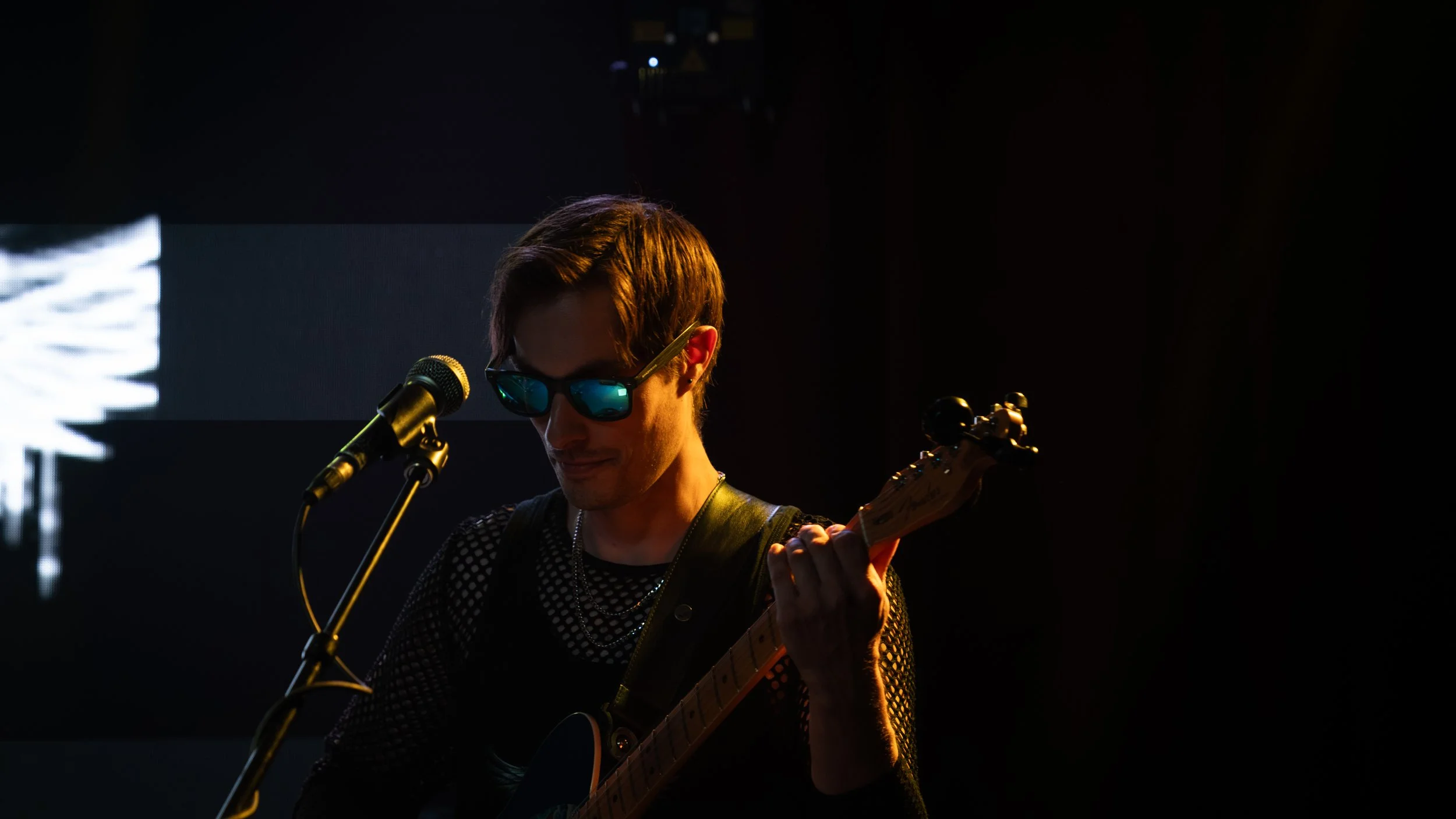A person with short hair wearing sunglasses and a mesh top playing an electric guitar on stage with minimal lighting.