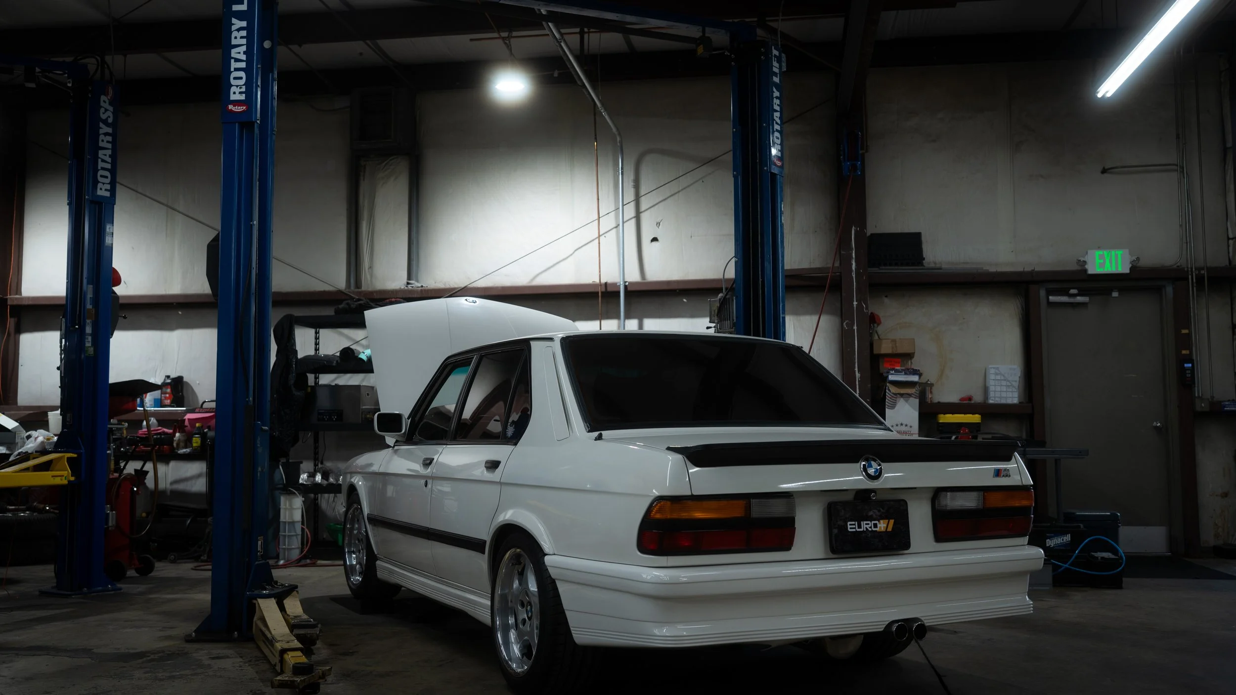 A white BMW E30 M3 parked inside a garage with a lift and various tools and equipment around.