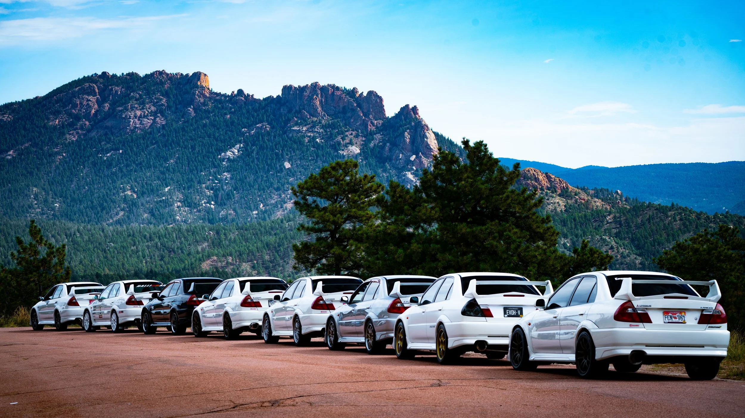 A parking lot filled with white cars with mountains and trees in the background under a blue sky.
