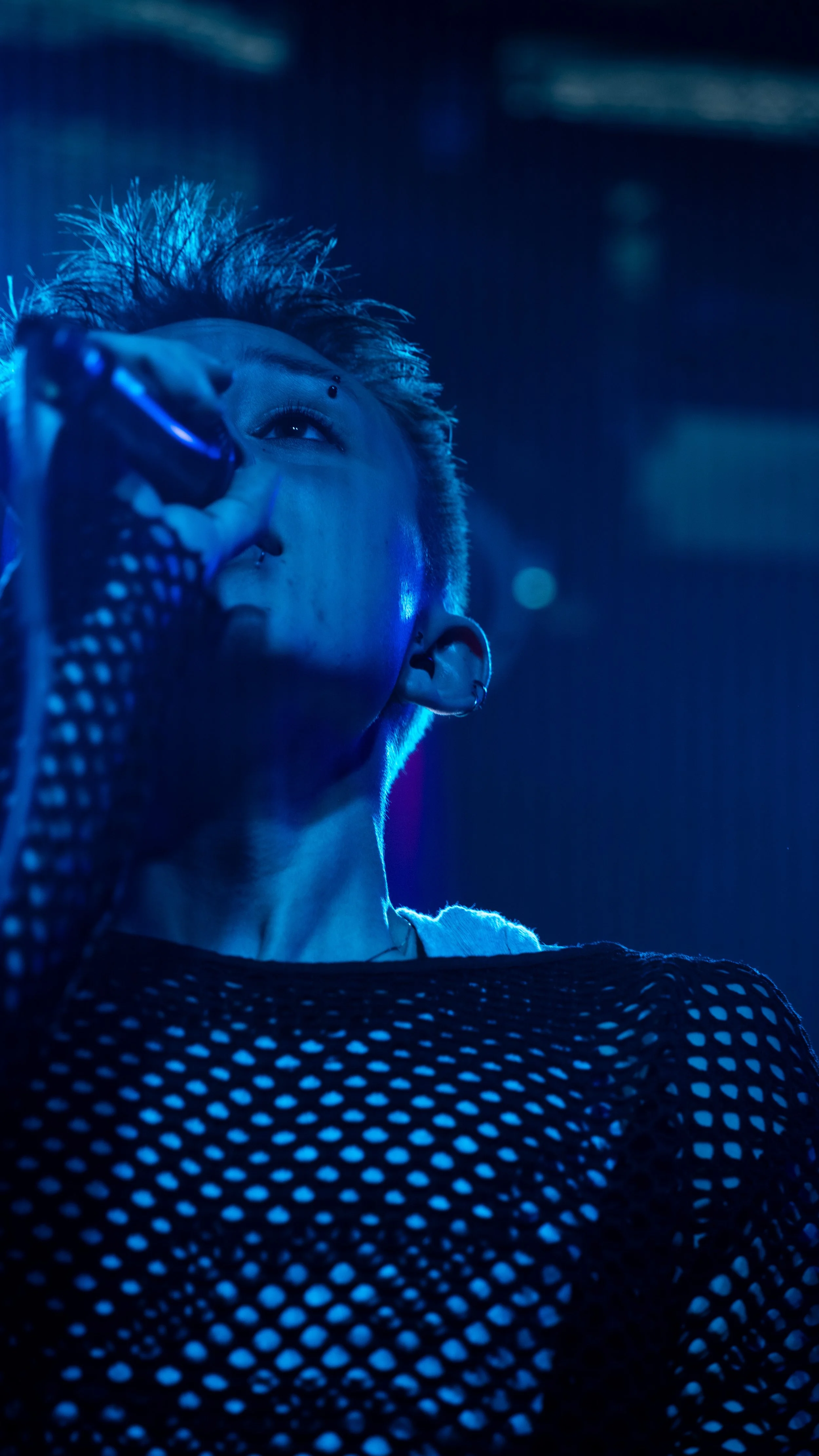 Person singing into a microphone on stage with blue lighting, wearing a black shirt with white polka dots.