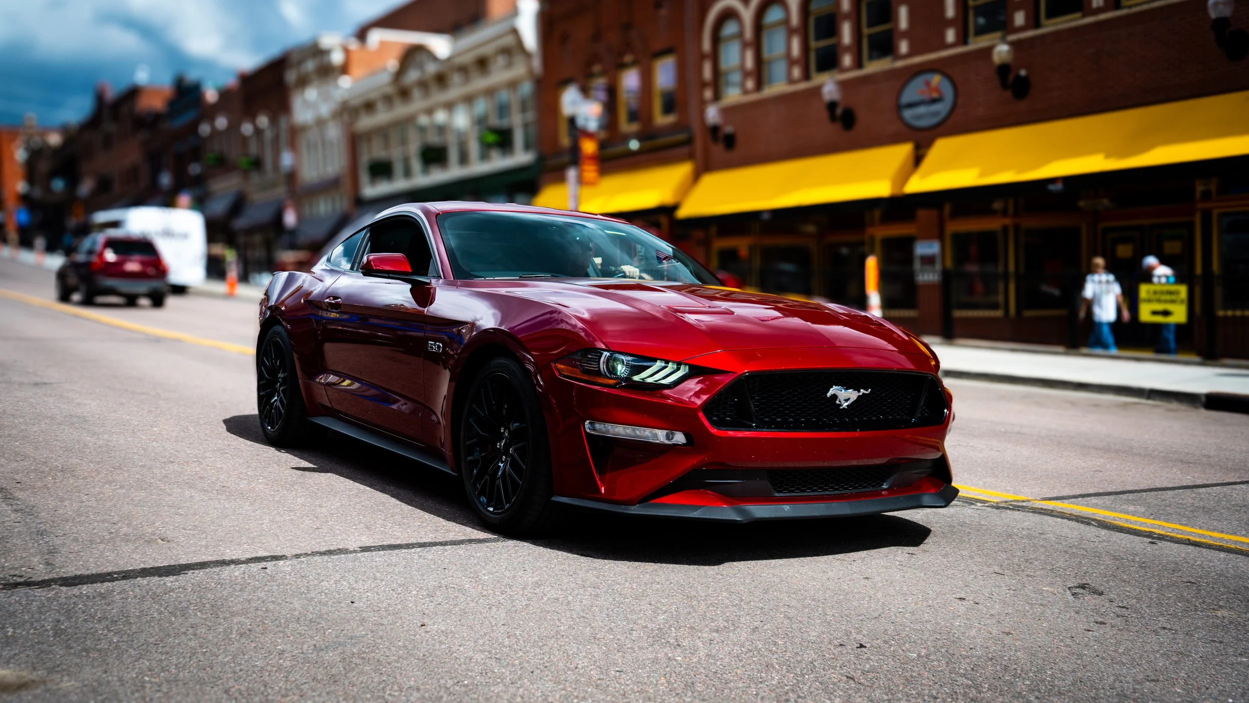 Red Ford Mustang sports car parked on a city street in front of storefronts with yellow awnings.