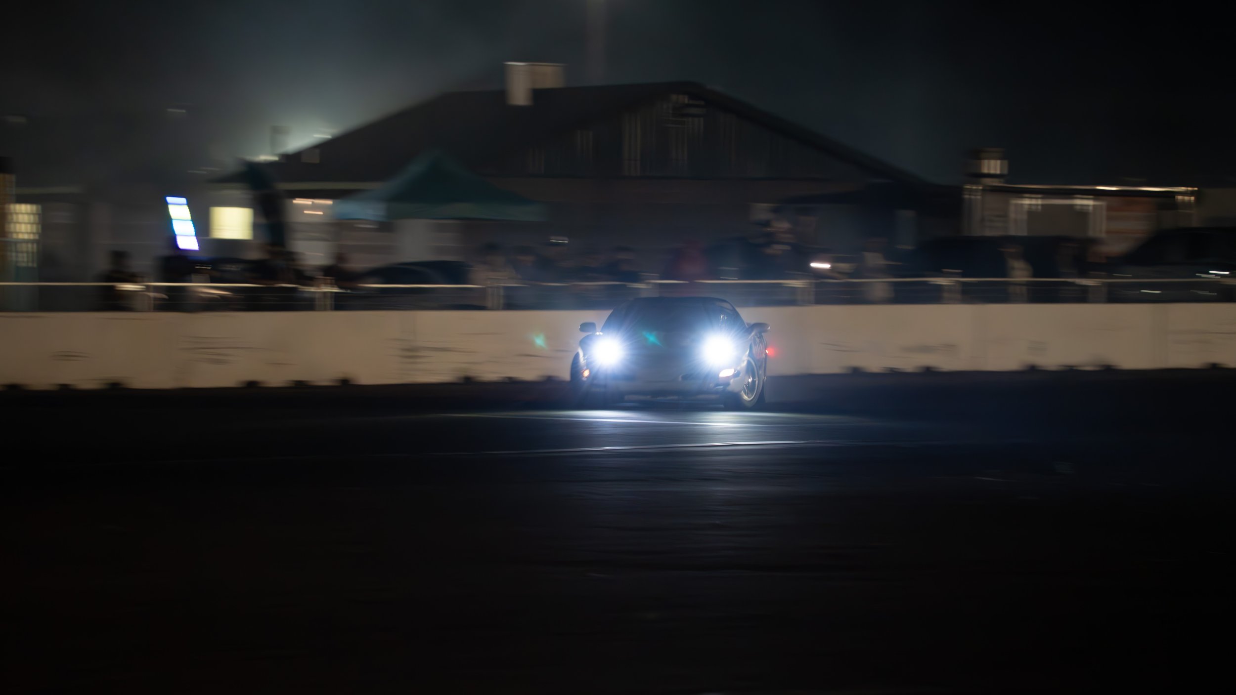 A black sports car with bright headlights driving on a race track at night, with a building and tents in the background.