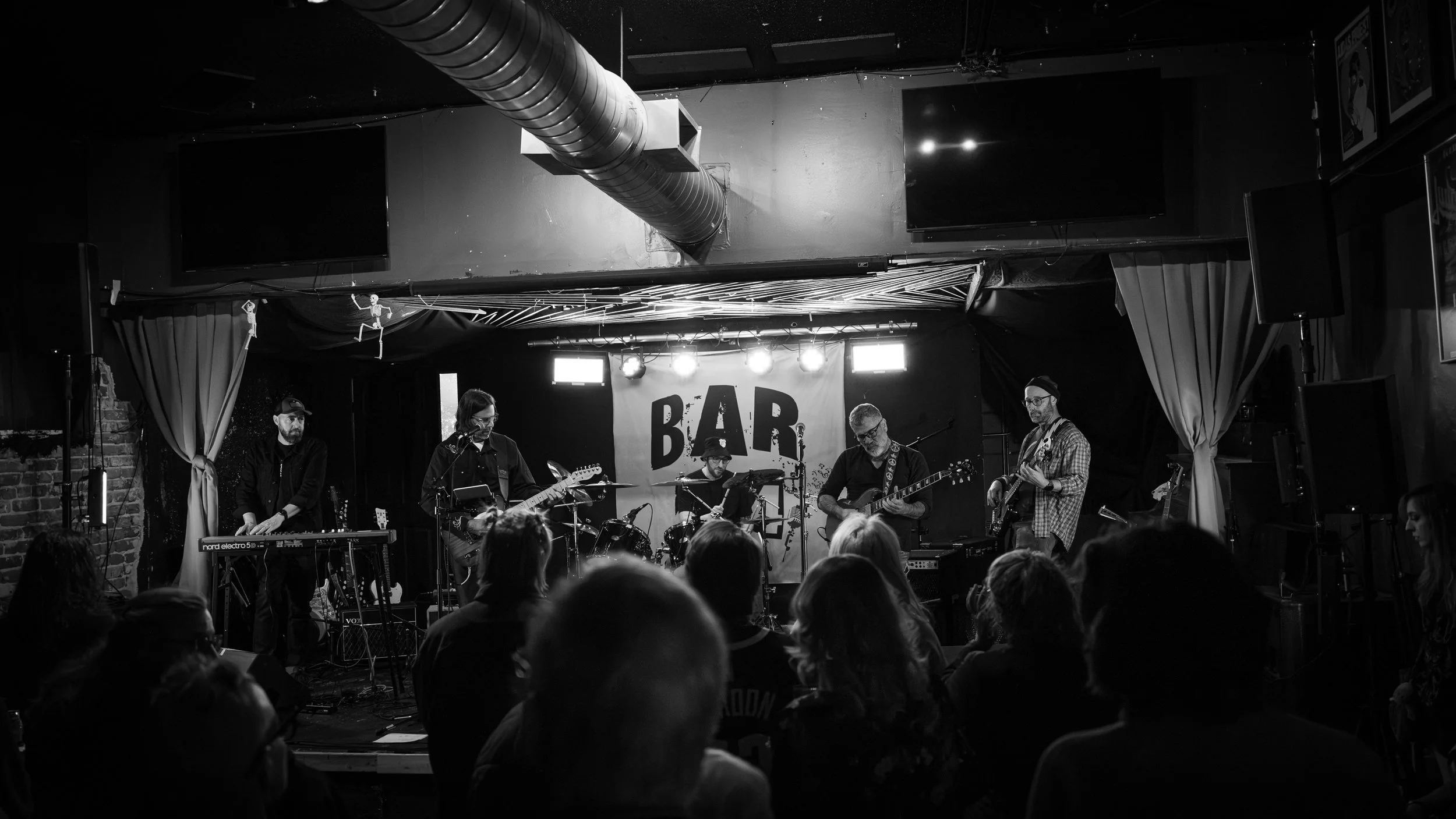 A live band performs on stage at a bar, with multiple musicians playing guitars, drums, and keyboards. The stage has a sign that says 'BAR' in the background, and the audience is watching in the foreground.
