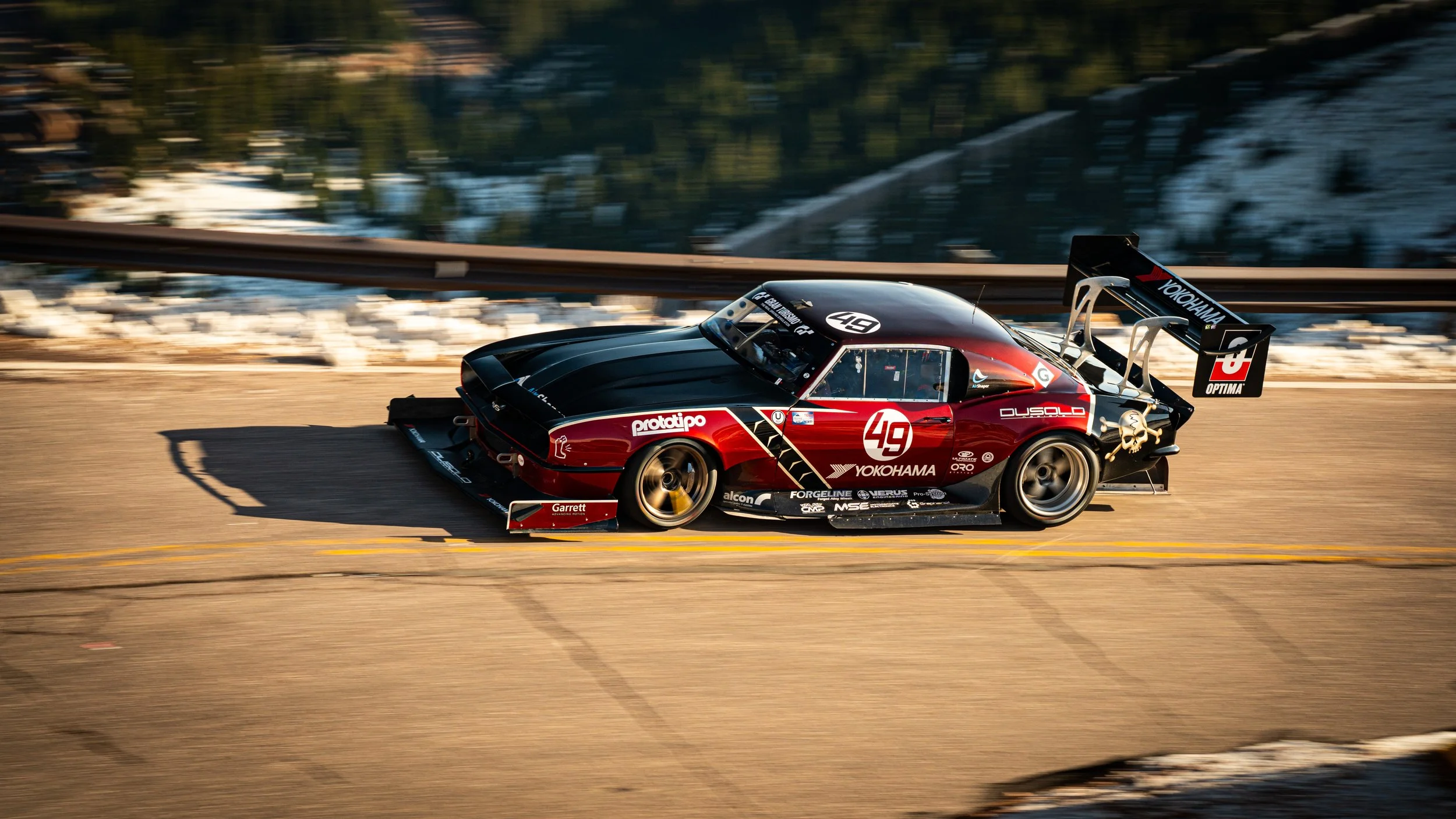 A race car driving on a mountain road with snow-capped mountains and trees in the background.