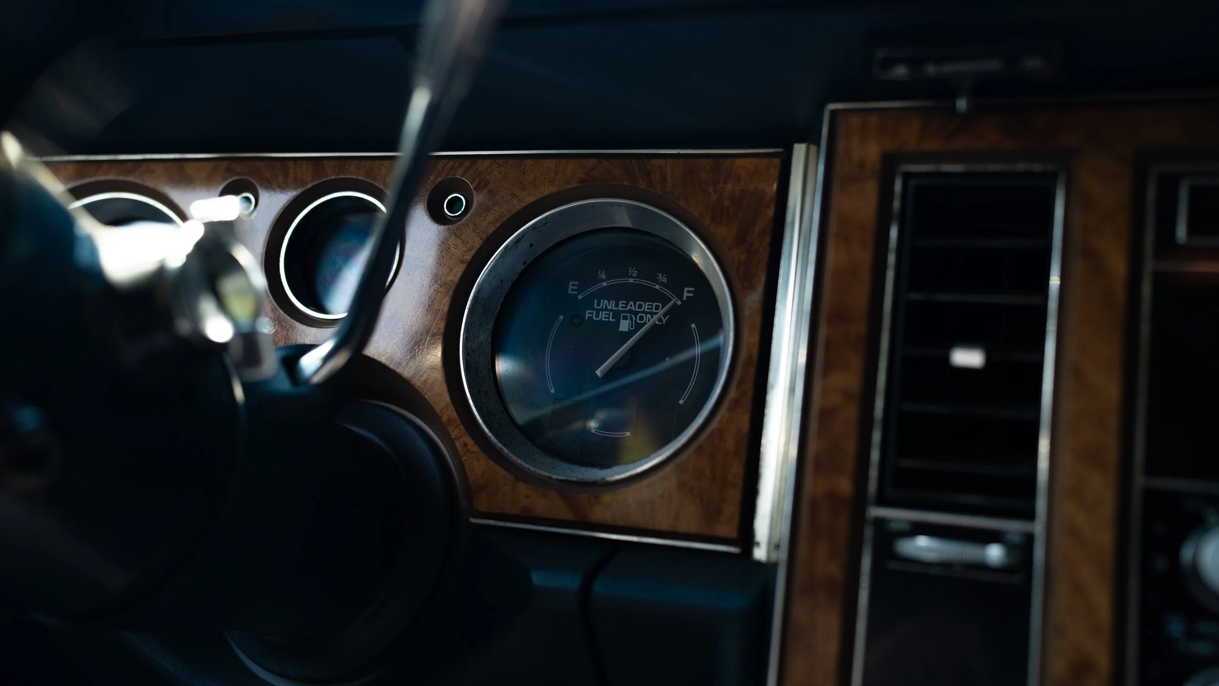 The image shows a close-up view of a car's dashboard, focusing on the fuel gauge which indicates the fuel level is near empty. The dashboard has a wood grain finish, and part of the steering wheel and air conditioning vents are visible.