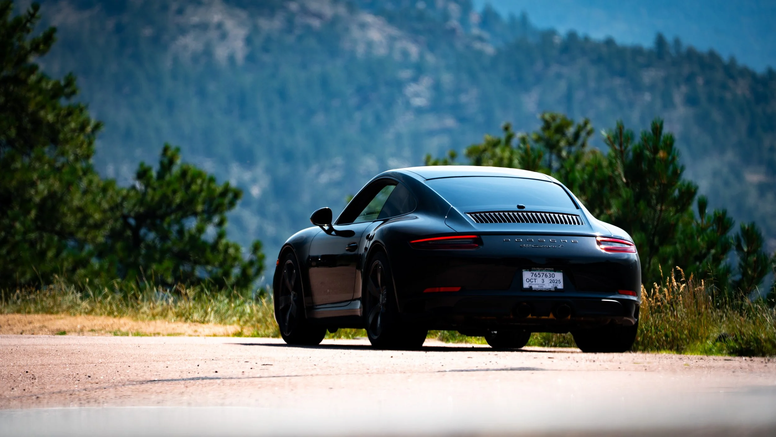 Black Porsche sports car parked outdoors with mountains and pine trees in the background.