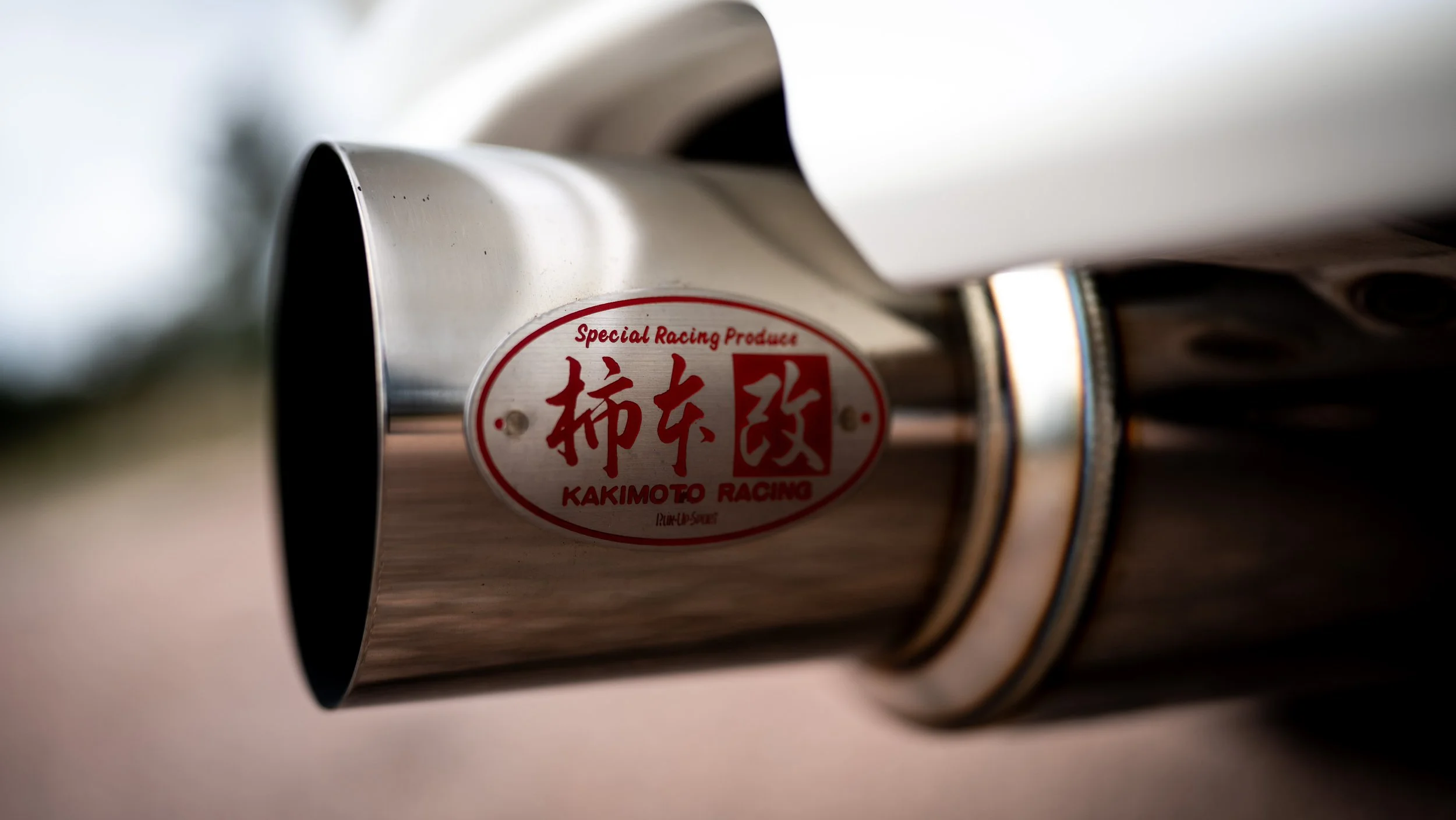 Close-up of a stainless steel car exhaust pipe with a red and white label that reads 'Special Racing Products, Kakimoto Racing'.