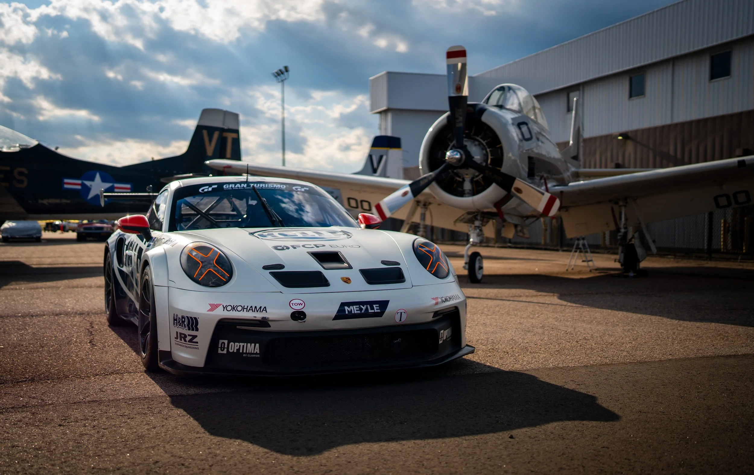 A white race car with sponsor logos parked on a tarmac surface, with a fighter jet and another airplane in the background, against a partly cloudy sky.