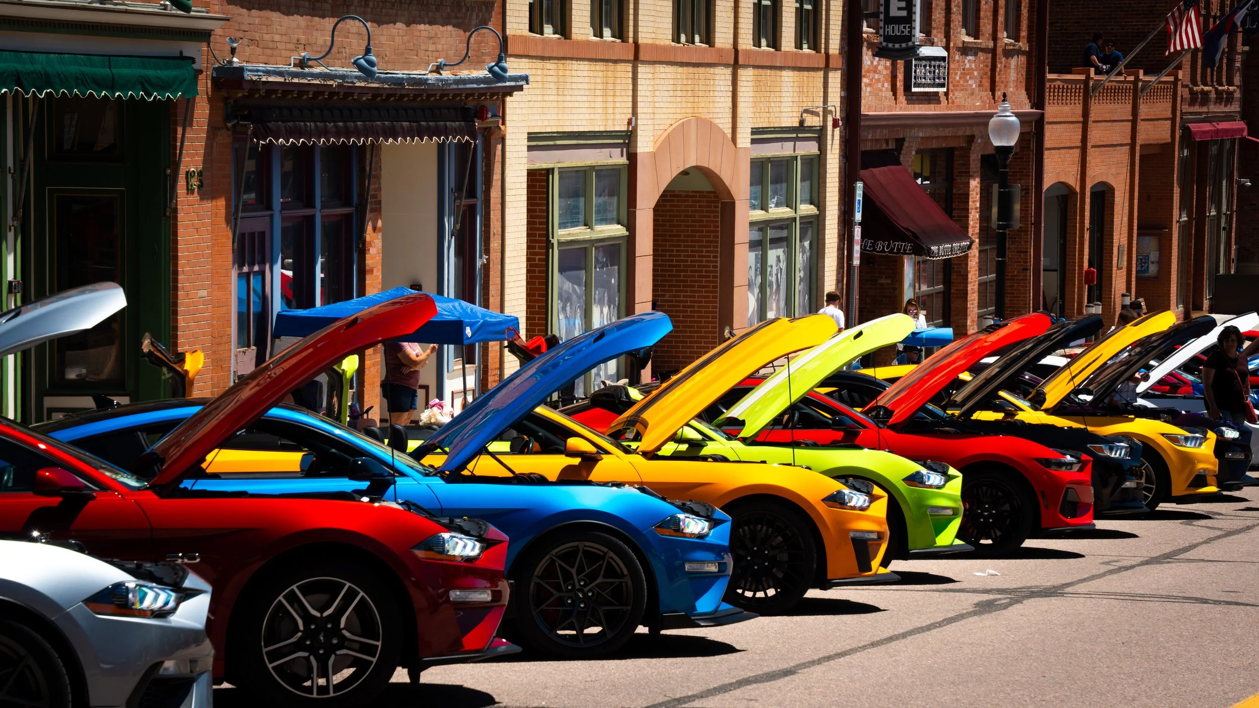 Colorful sports cars parked in a row with their hoods open on a city street, with brick buildings and pedestrians nearby.