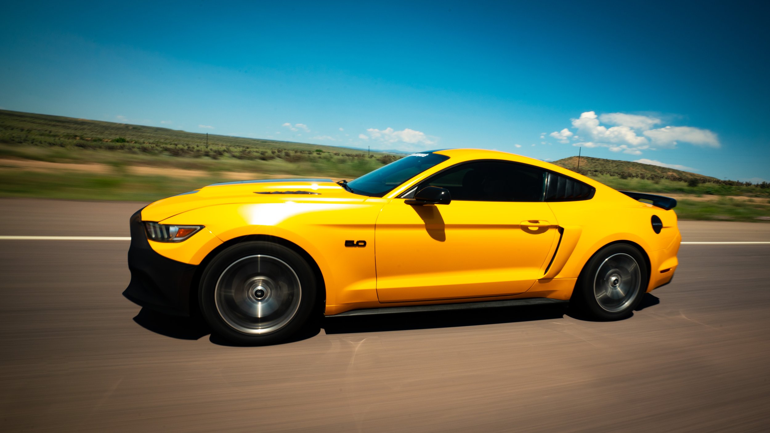 A yellow Ford Mustang GT driving on an open road with a background of green hills, clear blue sky, and scattered clouds.