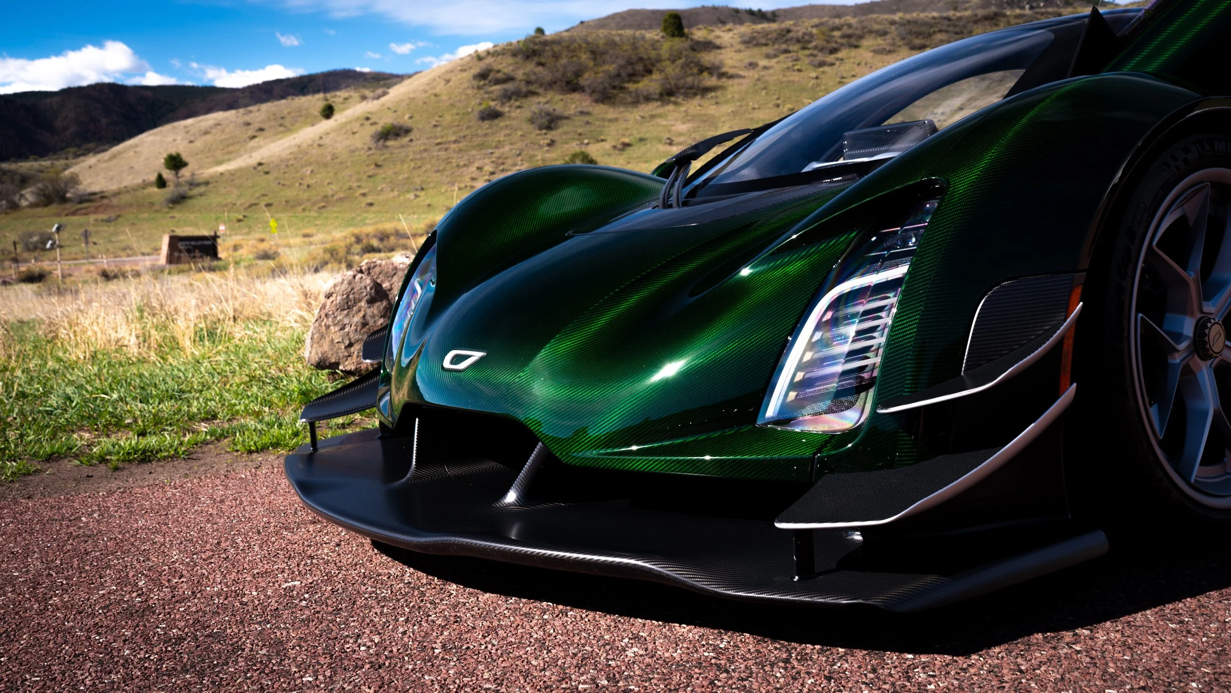 Close-up of a sleek green sports car with black accents parked on a red gravel path in a rural landscape with rolling hills, some trees, blue sky, and scattered clouds in the background.