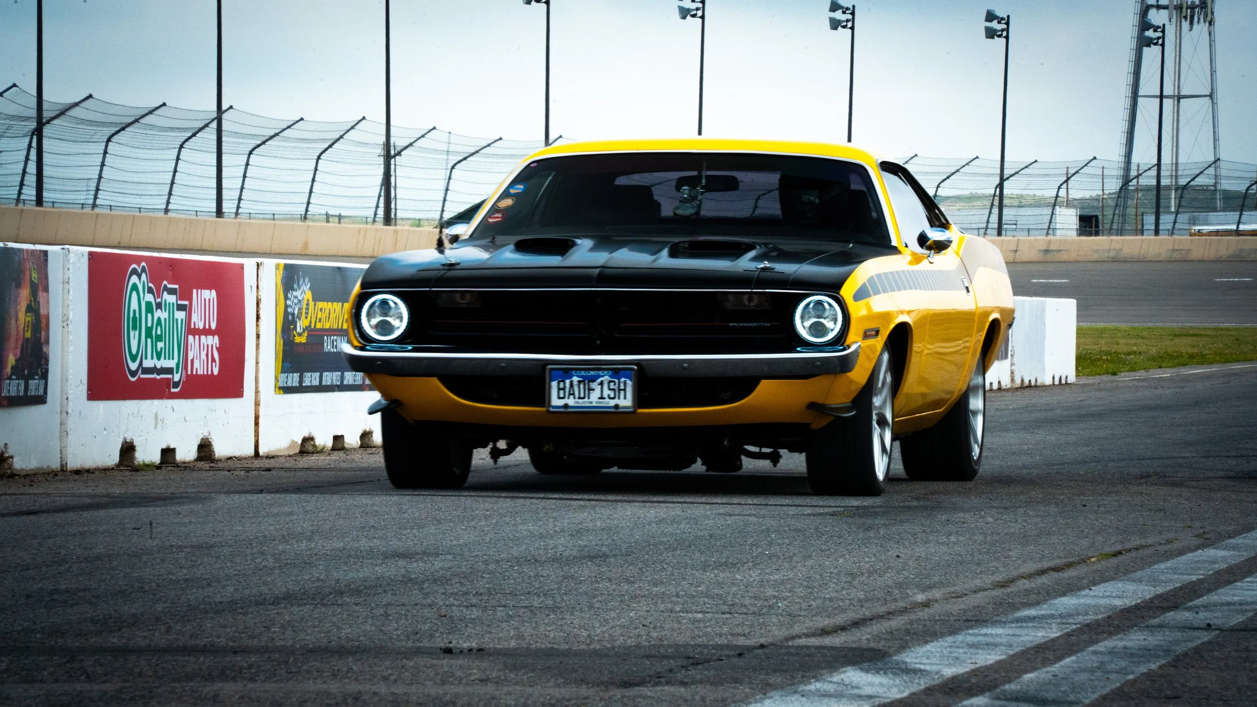 A yellow vintage muscle car on a race track, with a black hood and a license plate reading "BADFISH." The car is positioned next to a race track barrier with various advertisements, with a background of a fenced race track and tall light poles.