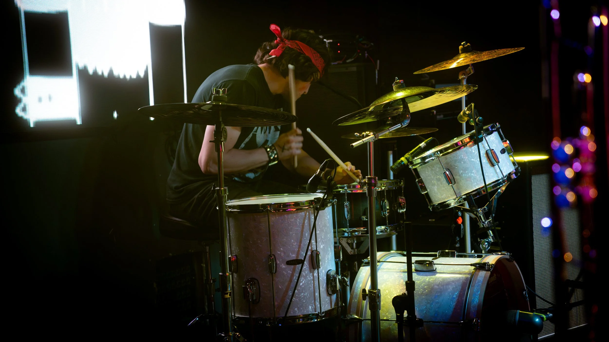 A person playing drums on stage, wearing a red bandana, with colorful stage lights and a large screen in the background.