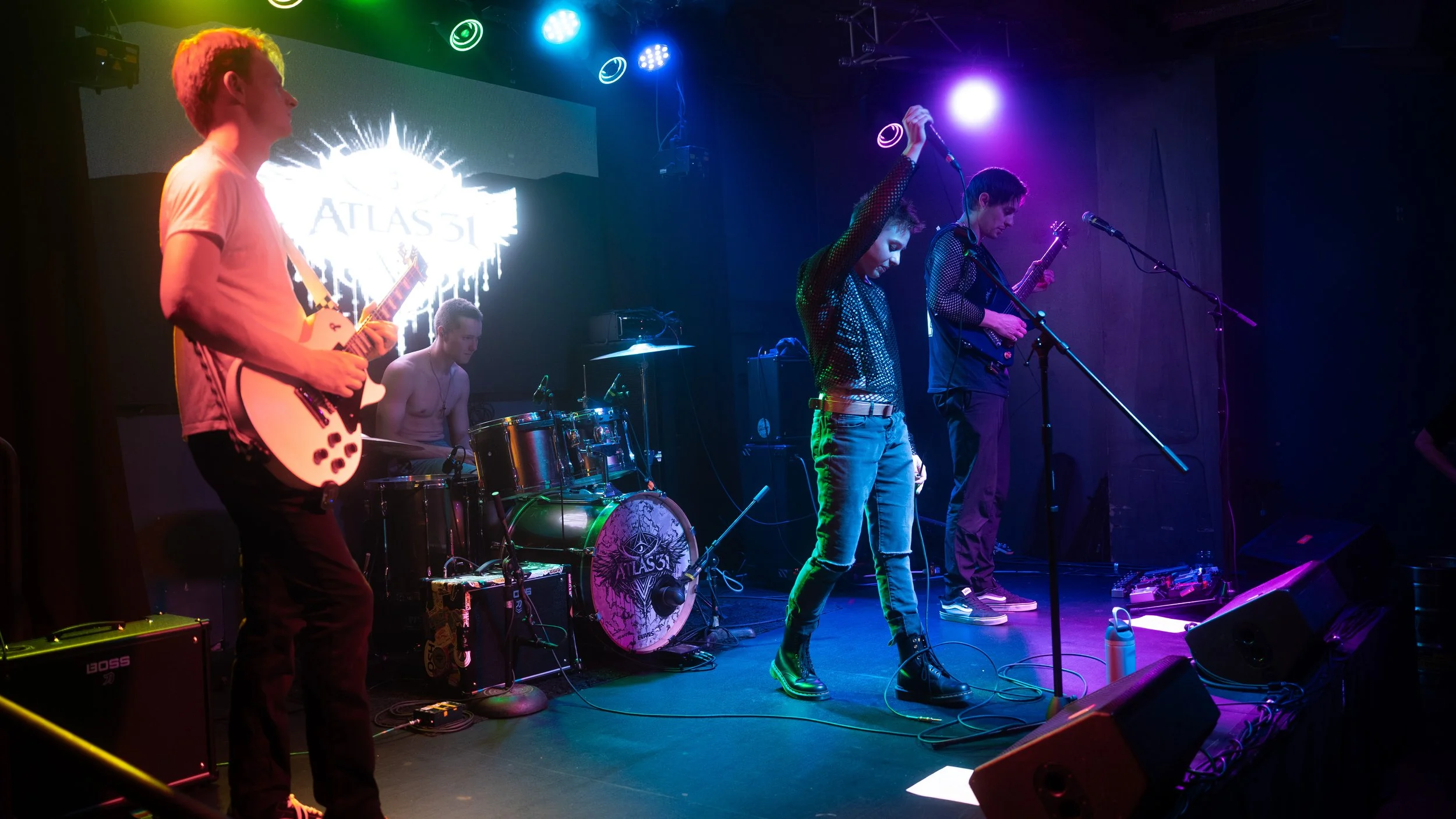 A band performing live on stage with colorful stage lights, including a guitarist, drummer, and singers, in a small venue. A large screen behind them displays the band's name, 'ATLAS.'