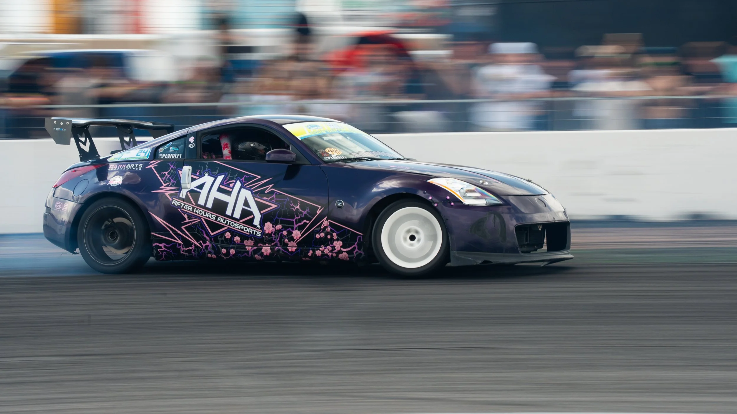 A black racing car with pink and purple graphics, including cherry blossoms and lightning bolts, speeding on a race track with a blurred crowd in the background.