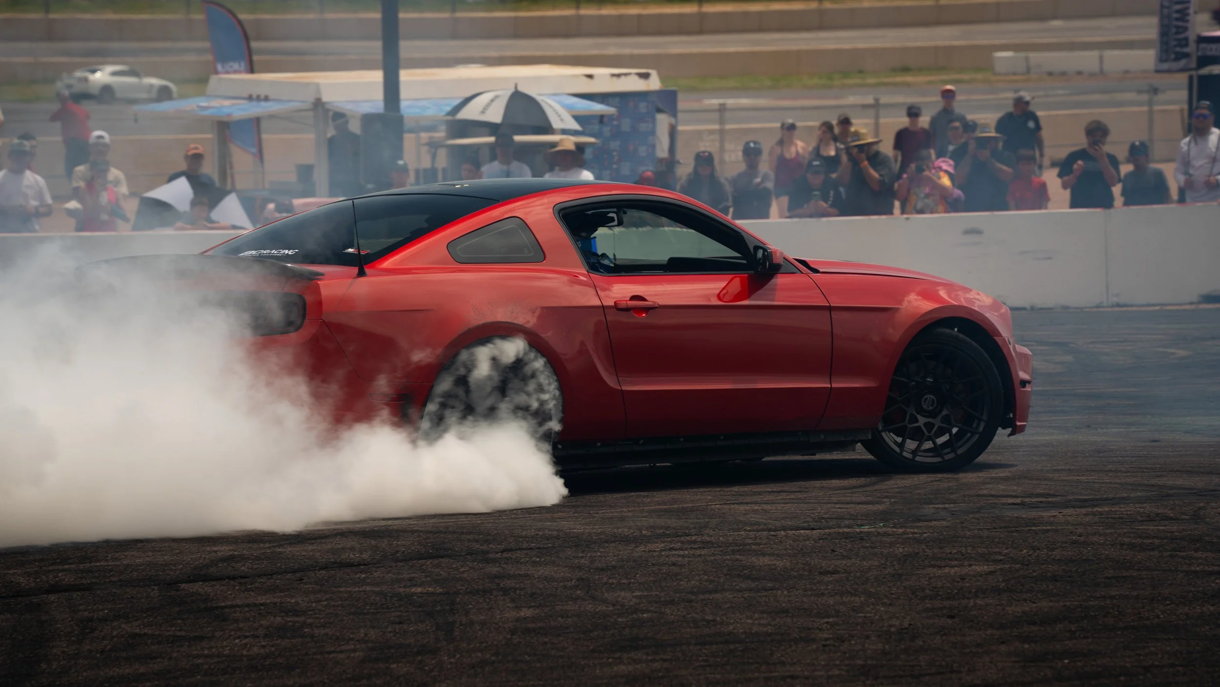 Red sports car performing a burnout during a racing event with spectators watching from behind the barriers.