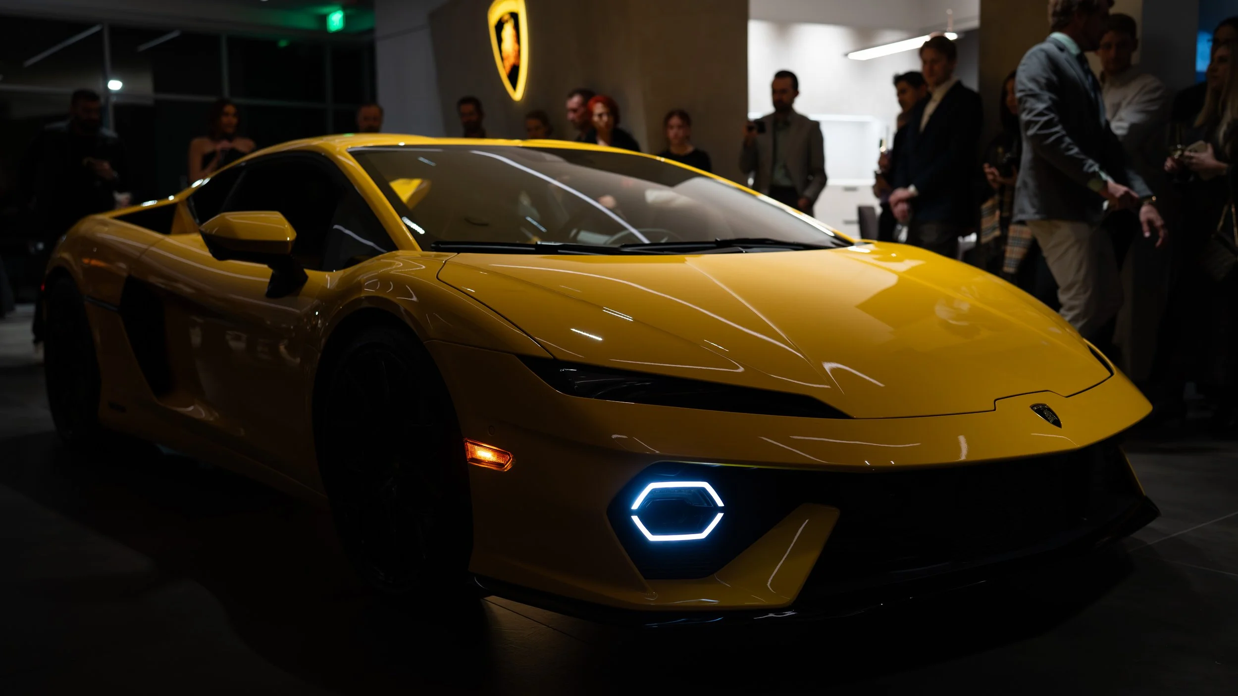 A yellow Lamborghini sports car displayed indoors at a car event, with people gathered around in the background.