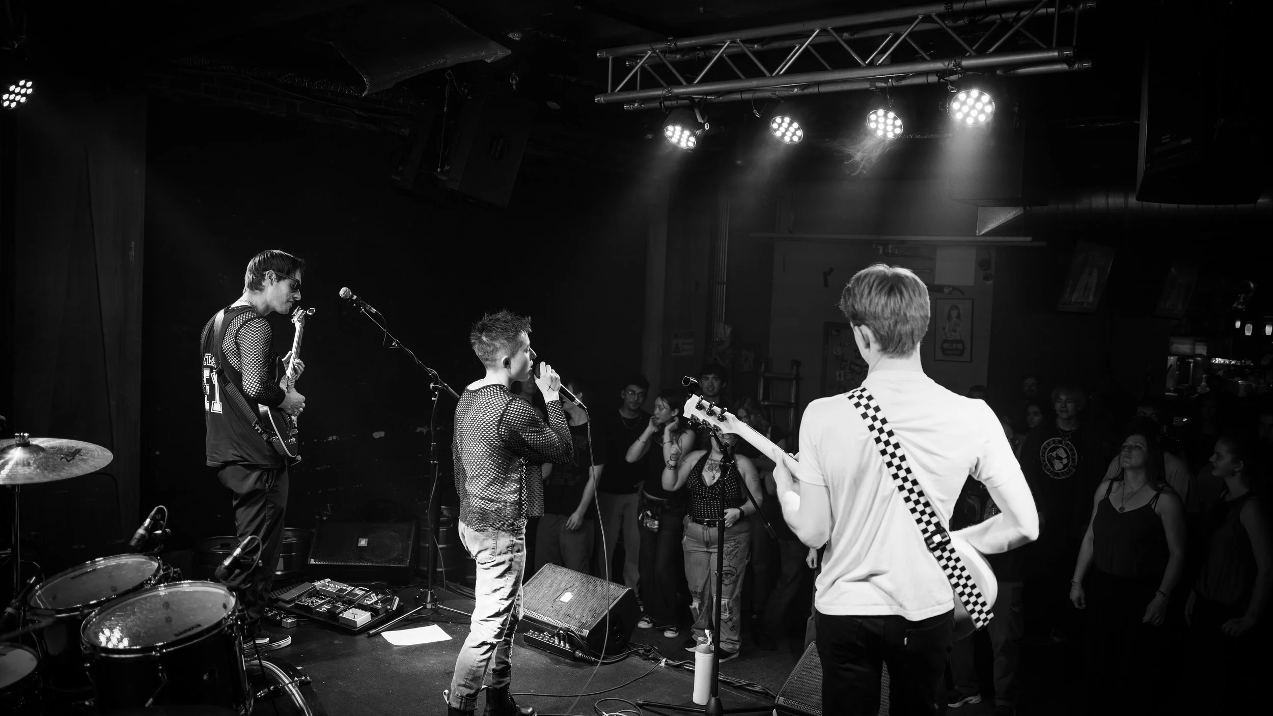 Black and white photo of a live music performance in a nightclub or bar, with a band playing on stage and audience members watching.