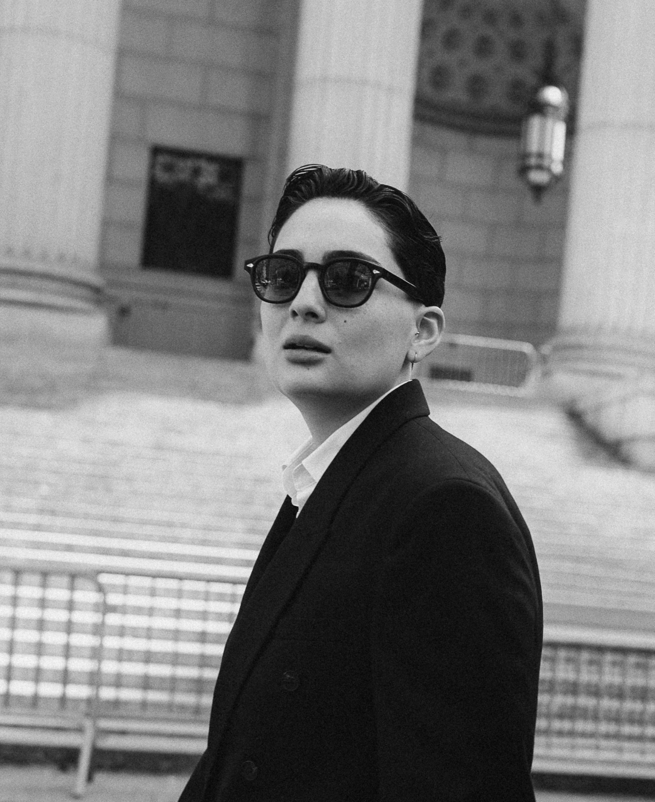 A black-and-white photo of a woman wearing sunglasses, a suit, and a blouse, standing outdoors with an urban background, including stairs, a building, and a lantern.