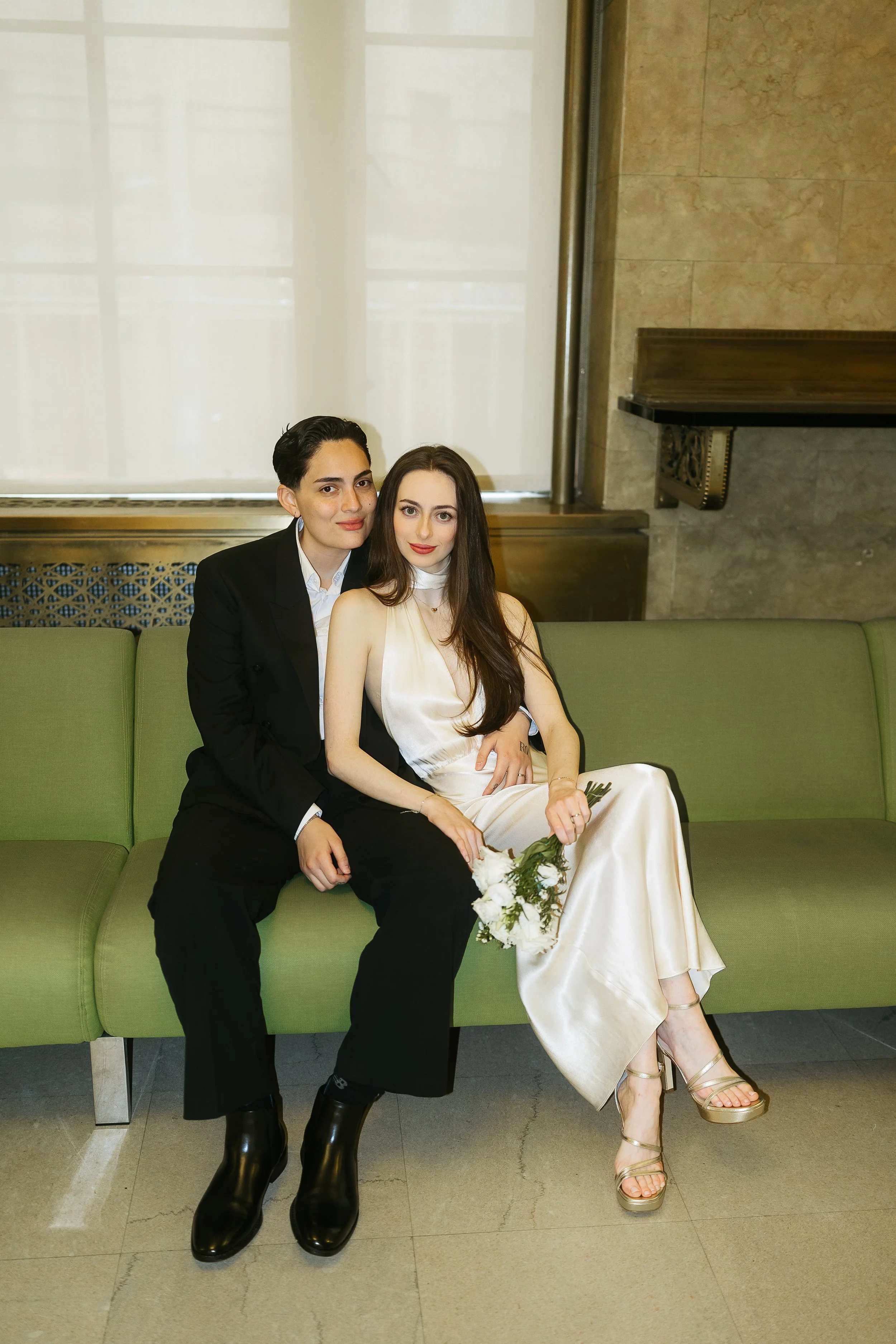 A happy couple holding a bridal bouquet while sitting on a green velvet sofa inside the Manhattan Marriage Bureau, waiting for their NYC courthouse wedding ceremony.