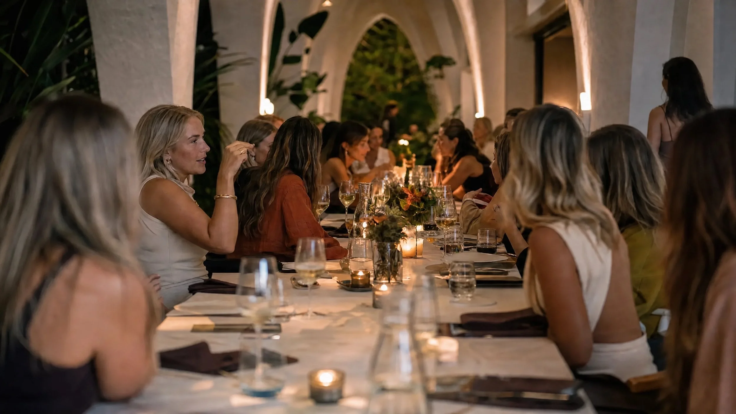 Women gathered around a long dining table at an evening gathering, engaged in conversation, with candles and a floral centerpiece on the table, in a well-lit outdoor or semi-outdoor venue with arches.