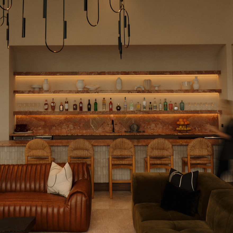 A modern bar area with a marble countertop, five wooden chairs, and a backlit shelf holding various bottles and decorative items. There are two sofas in front, one brown leather with white and black pillows, and one green with black pillows. A person