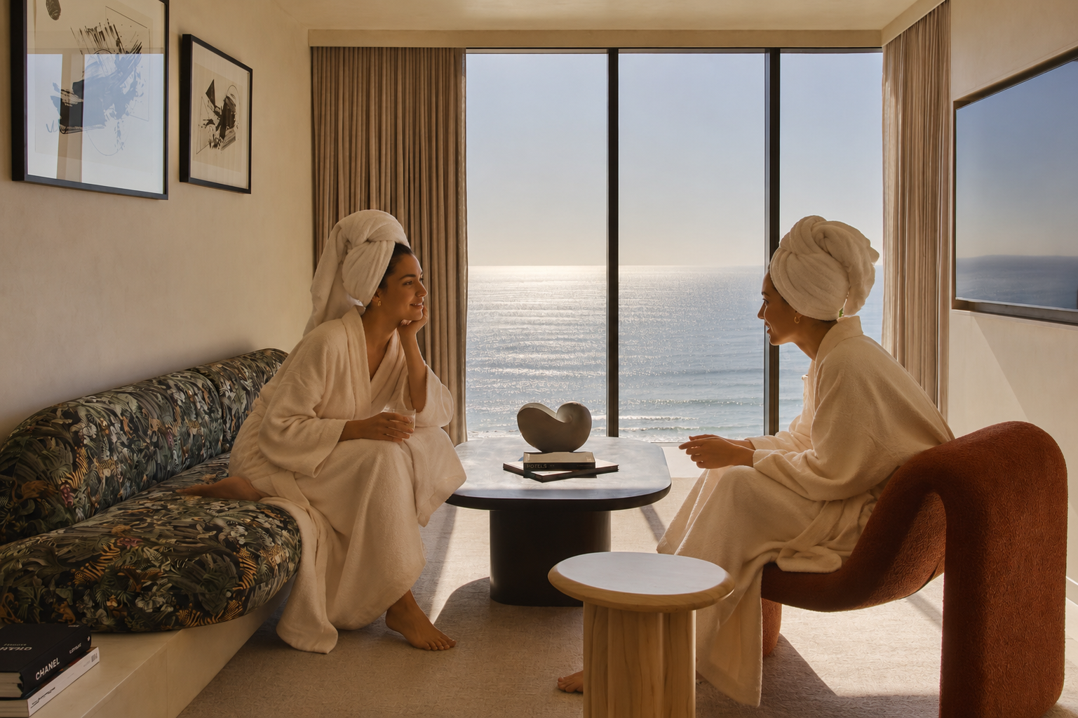 Two women in bathrobes with towels wrapped around their heads having a conversation in a hotel room with ocean view, sitting across from each other near a coffee table with a heart-shaped sculpture and books.