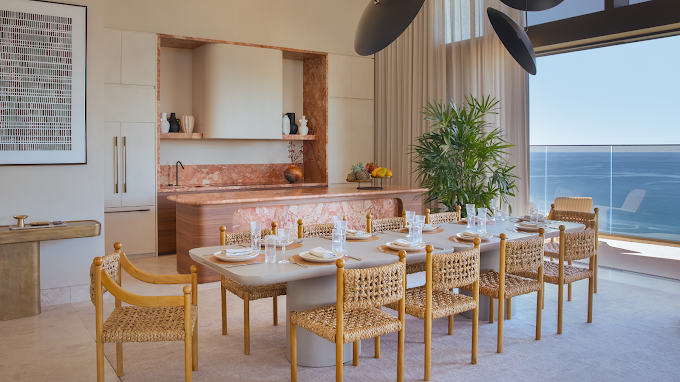 Luxury dining room with a long white table, eight wicker chairs, table setting, large window with ocean view, potted plant, and modern decor.