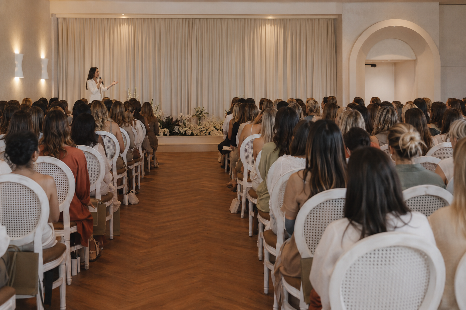 Woman speaking at the front of a large room filled with seated women.