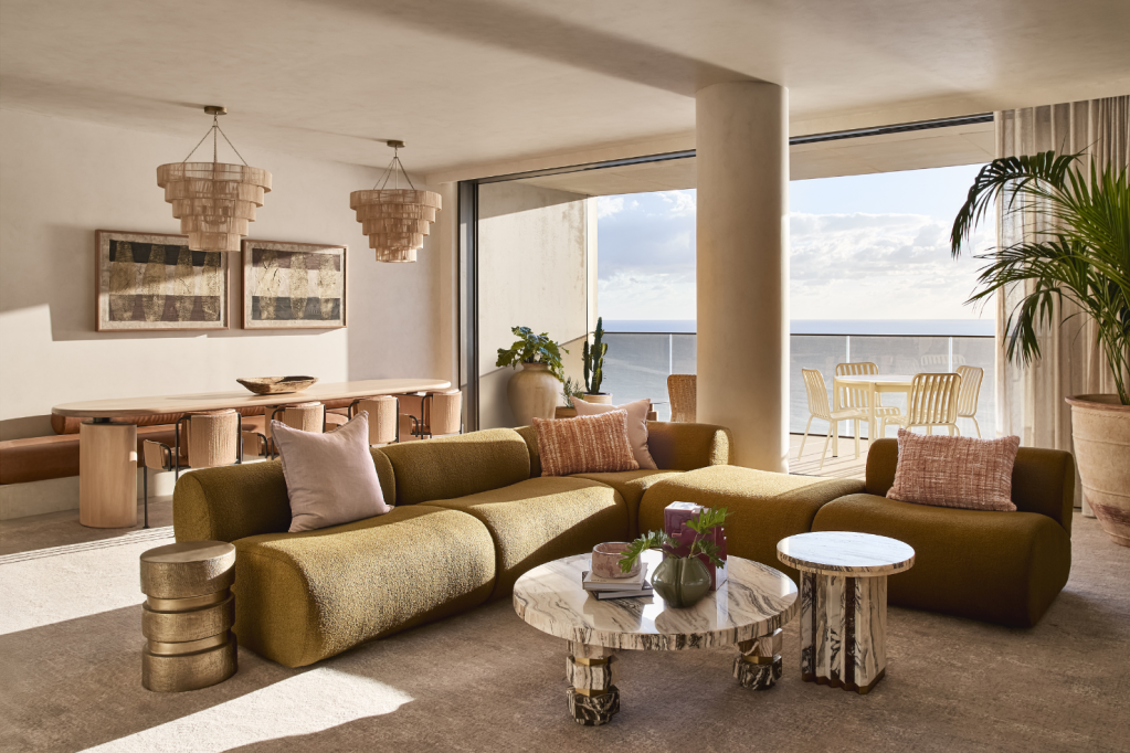 Modern living room with yellow sectional sofa, marble coffee tables, and large potted plants, overlooking an outdoor balcony with ocean view.