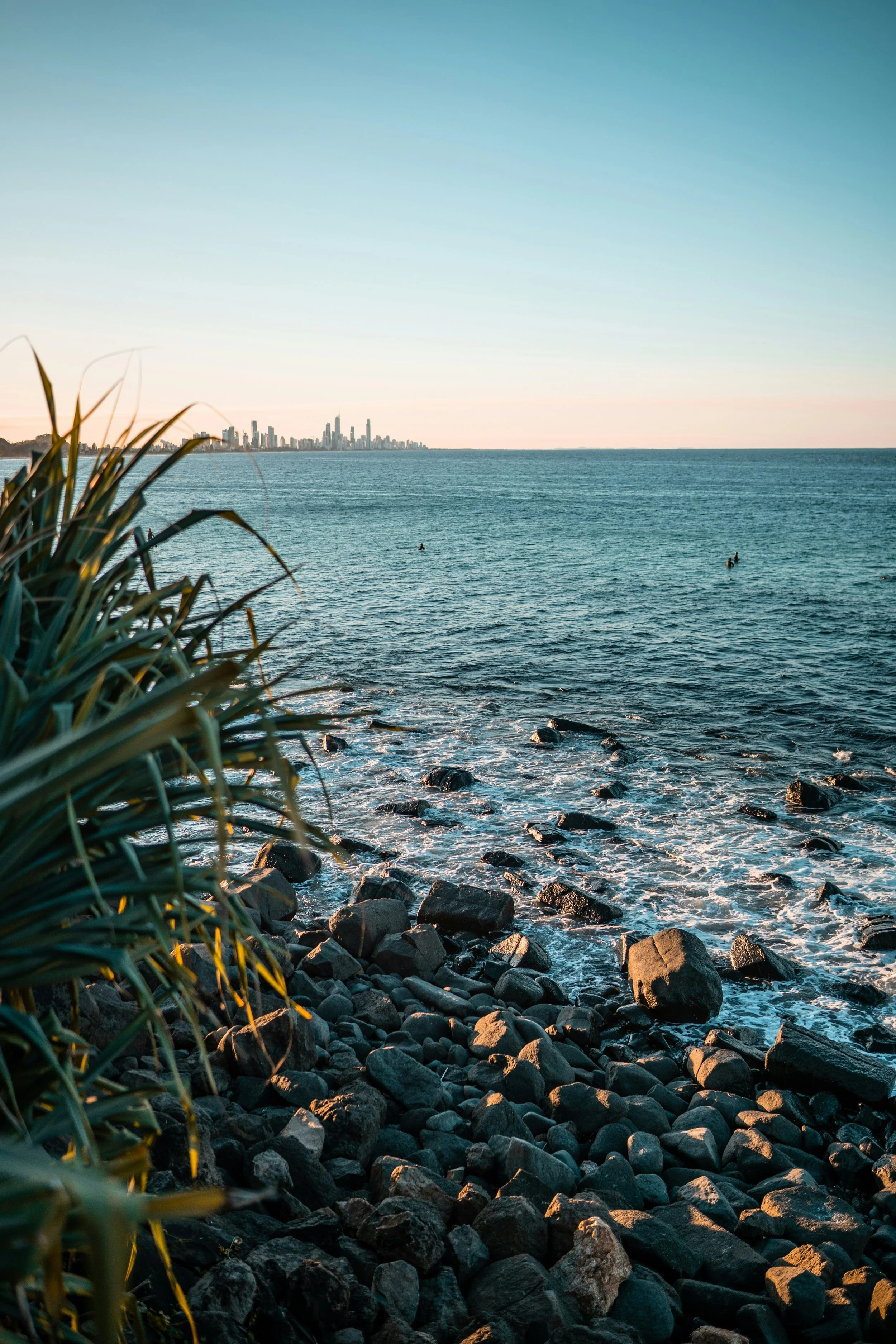 View of the ocean with waves hitting rocks along the shoreline, distant city skyline visible under a clear sky during sunset.