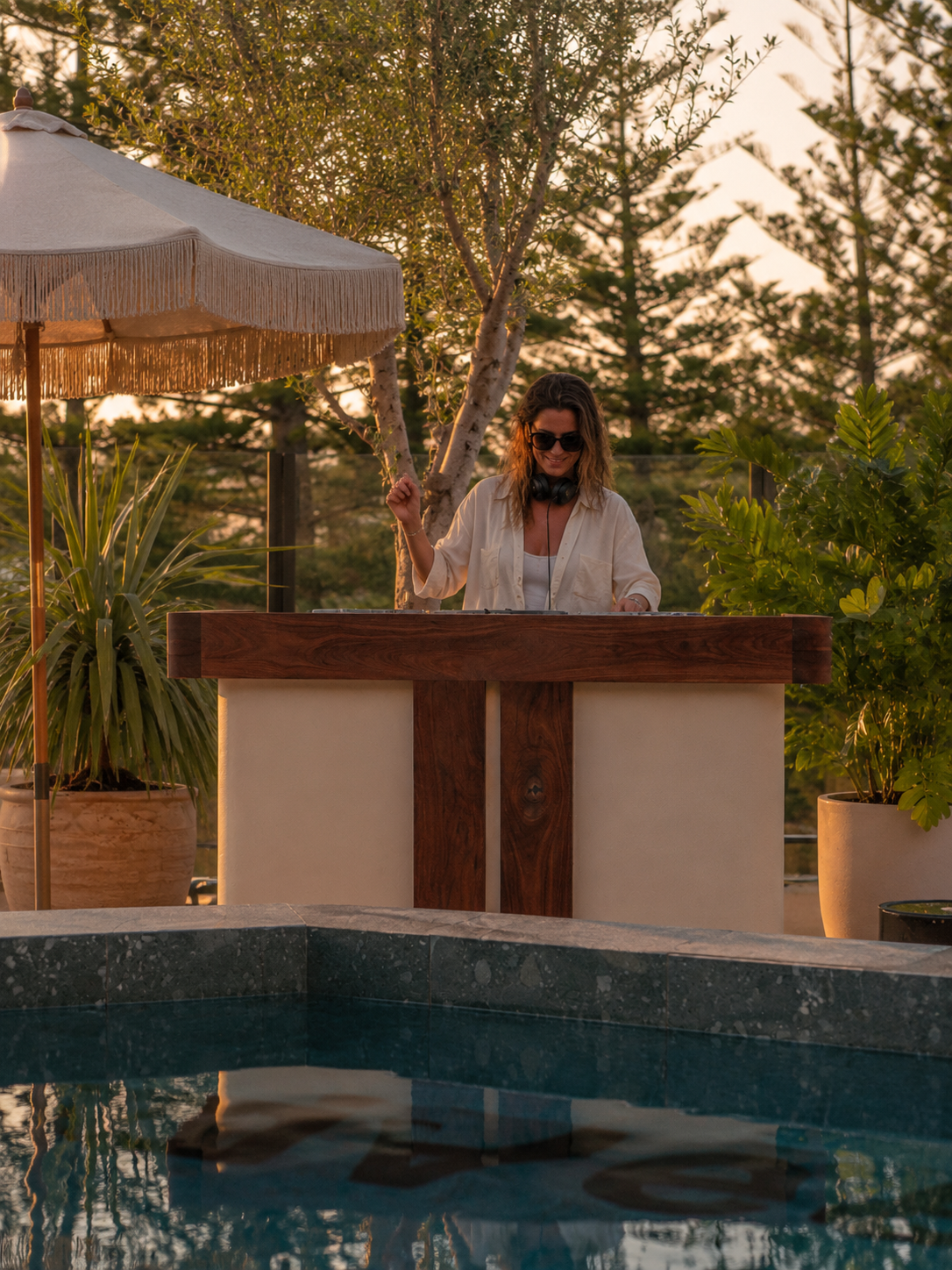 A woman with sunglasses and headphones DJing outdoors by a swimming pool during sunset, with trees and greenery in the background.