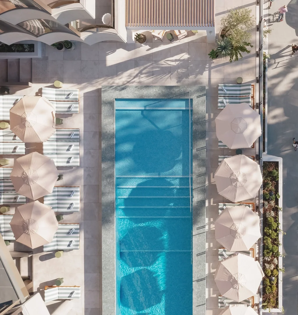 An aerial view of a hotel pool area with lounge chairs and umbrellas. The rectangular swimming pool is in the center, surrounded by striped lounge chairs on both sides. There are beige umbrellas over the lounge chairs, with some patio furniture and plants on the nearby patio area.