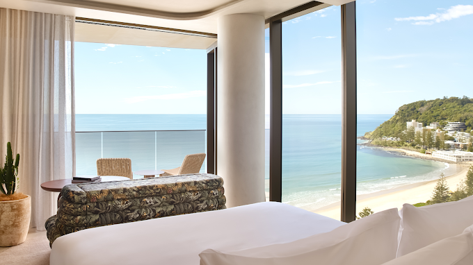 Luxury hotel room with large floor-to-ceiling windows overlooking the beach and ocean, with two chairs and a small table on a balcony.