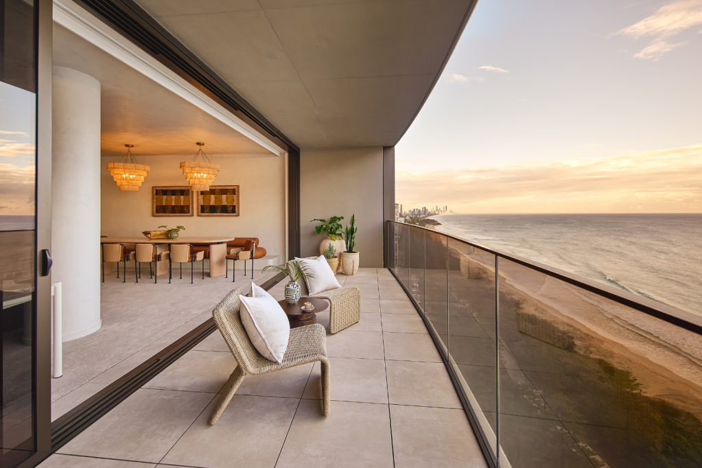 Balcony with two chairs and pillows overlooking ocean, with dining area inside visible through sliding door.
