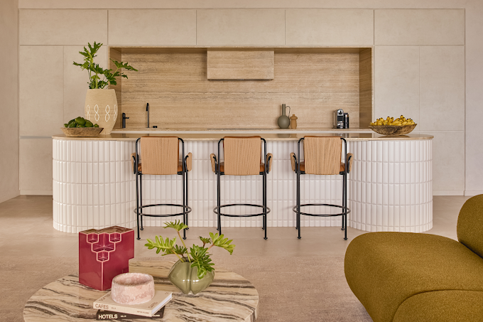 Modern kitchen with a white curved island counter, three chairs with beige cushions and black frames, and decorative items including a plant, vases, and a bowl of lemons.