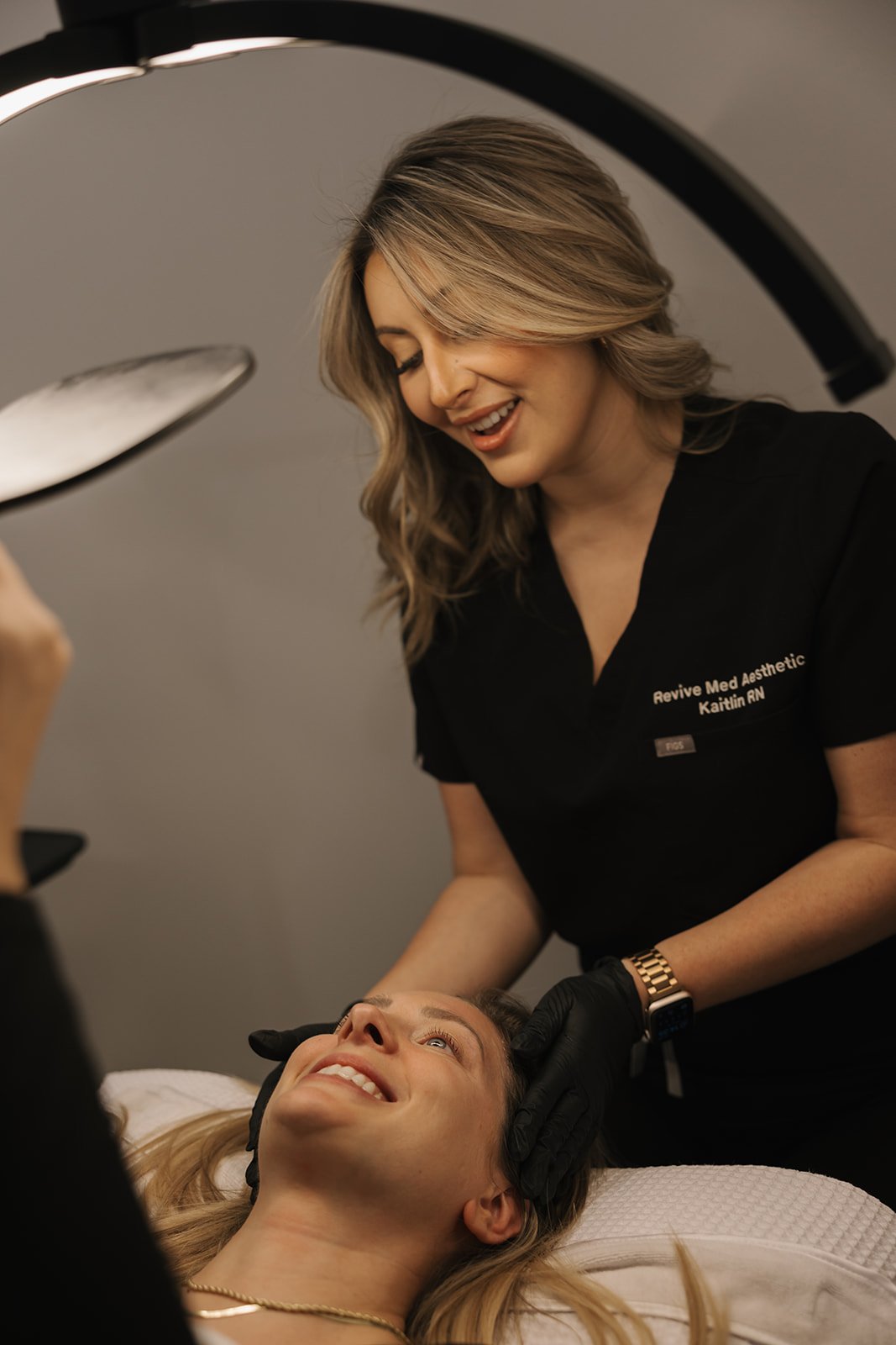 A woman lying on an examination table receiving a cosmetic facial treatment from a medical aesthetic professional in black scrubs.