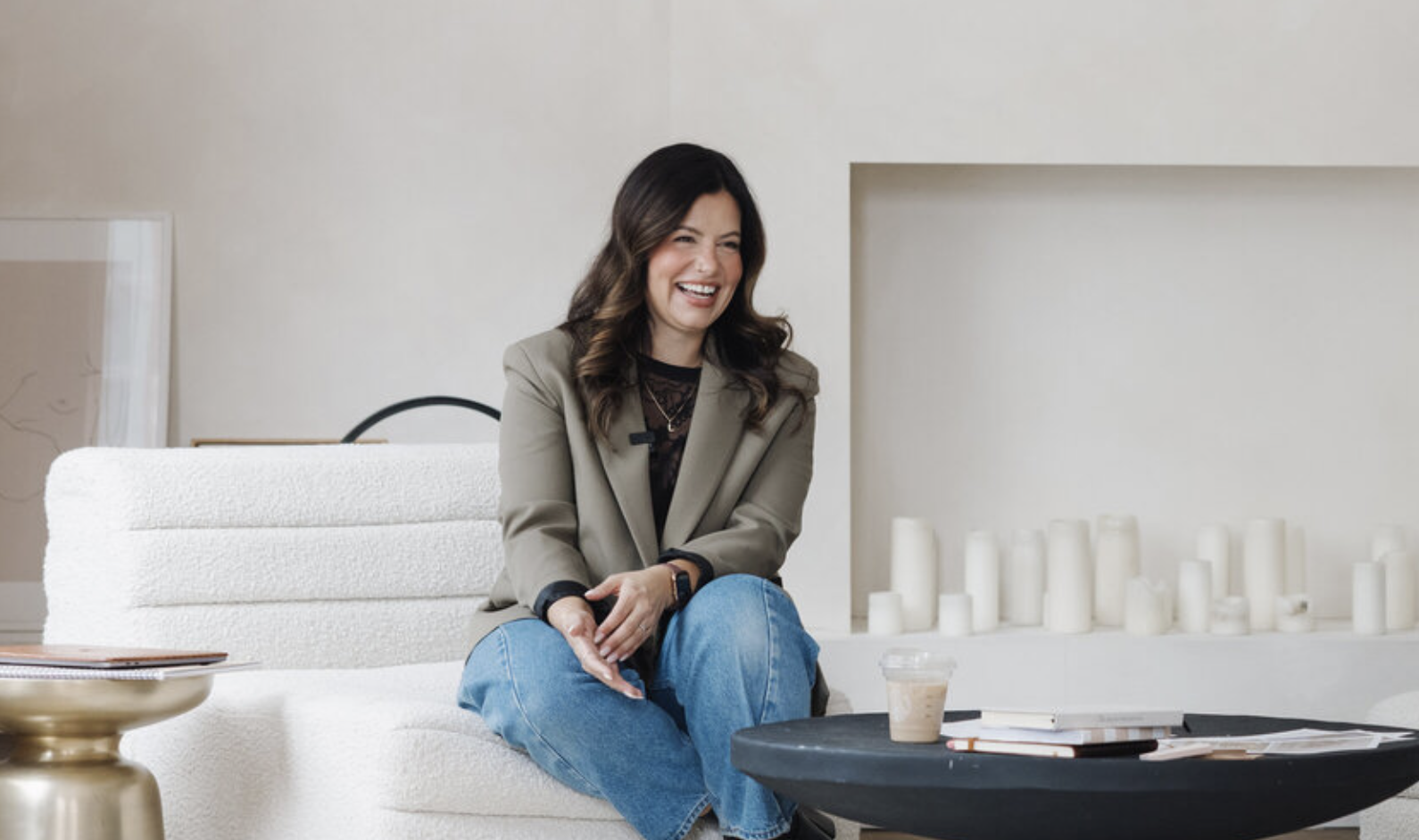 Woman sitting on a white sofa, smiling and laughing, with a coffee table in front of her containing a plastic cup and some books, in a bright, minimalist room.