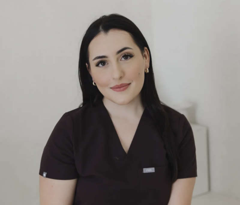 Portrait of a woman with long dark hair, wearing a dark medical scrub top, smiling softly, in a clinical setting.