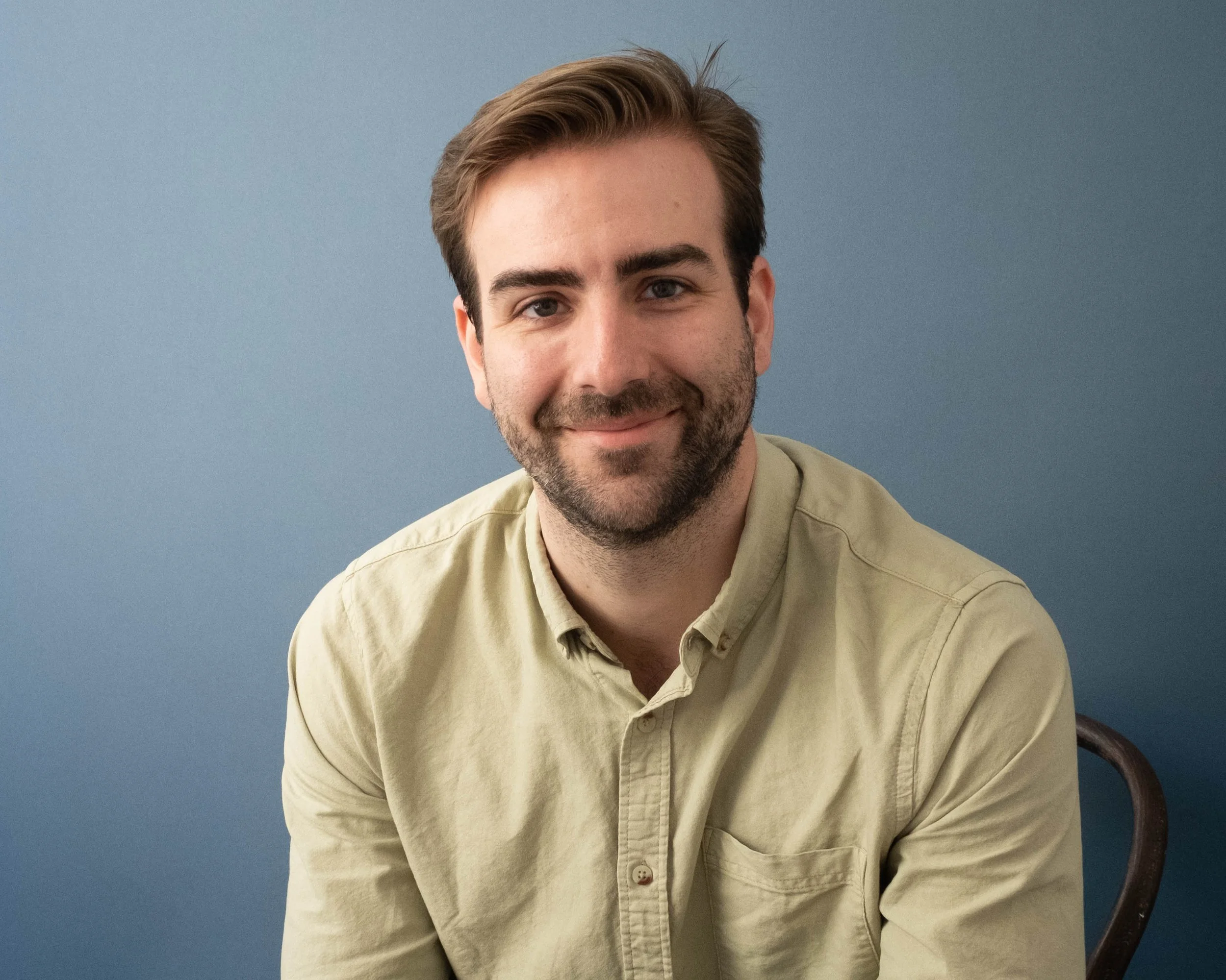 A man with dark blonde hair, blue eyes, and a beard sitting in front of a blue background, smiling, wearing a light beige shirt. Nickolas Hasse, documentary producer.