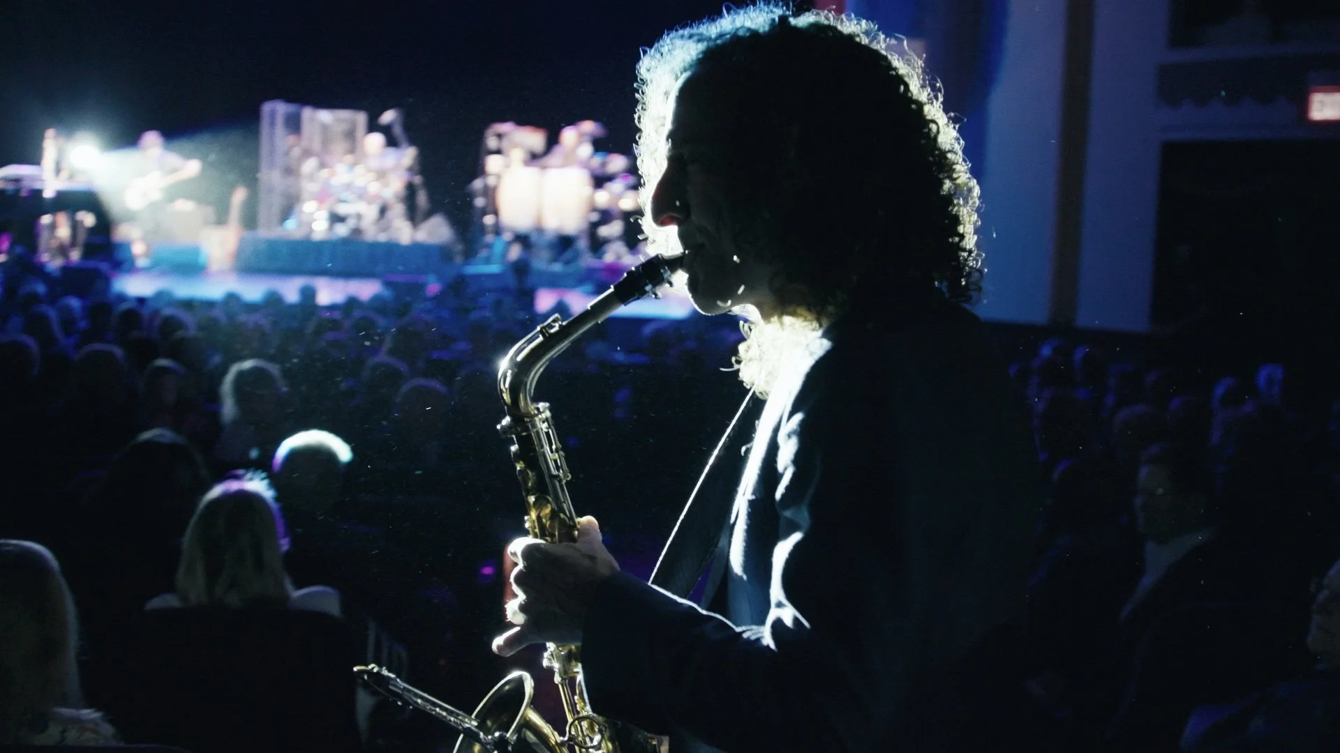 A person playing the saxophone during a live concert, with a large crowd and stage with lights and musicians in the background. Kenny G documentary.