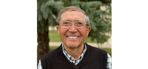 A smiling elderly man wearing glasses, a plaid shirt, and a black sweater vest outdoors near a pine tree.