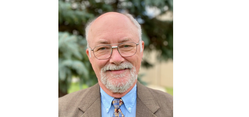A senior man with glasses, a bald head, and a white beard, smiling outdoors with trees in the background.